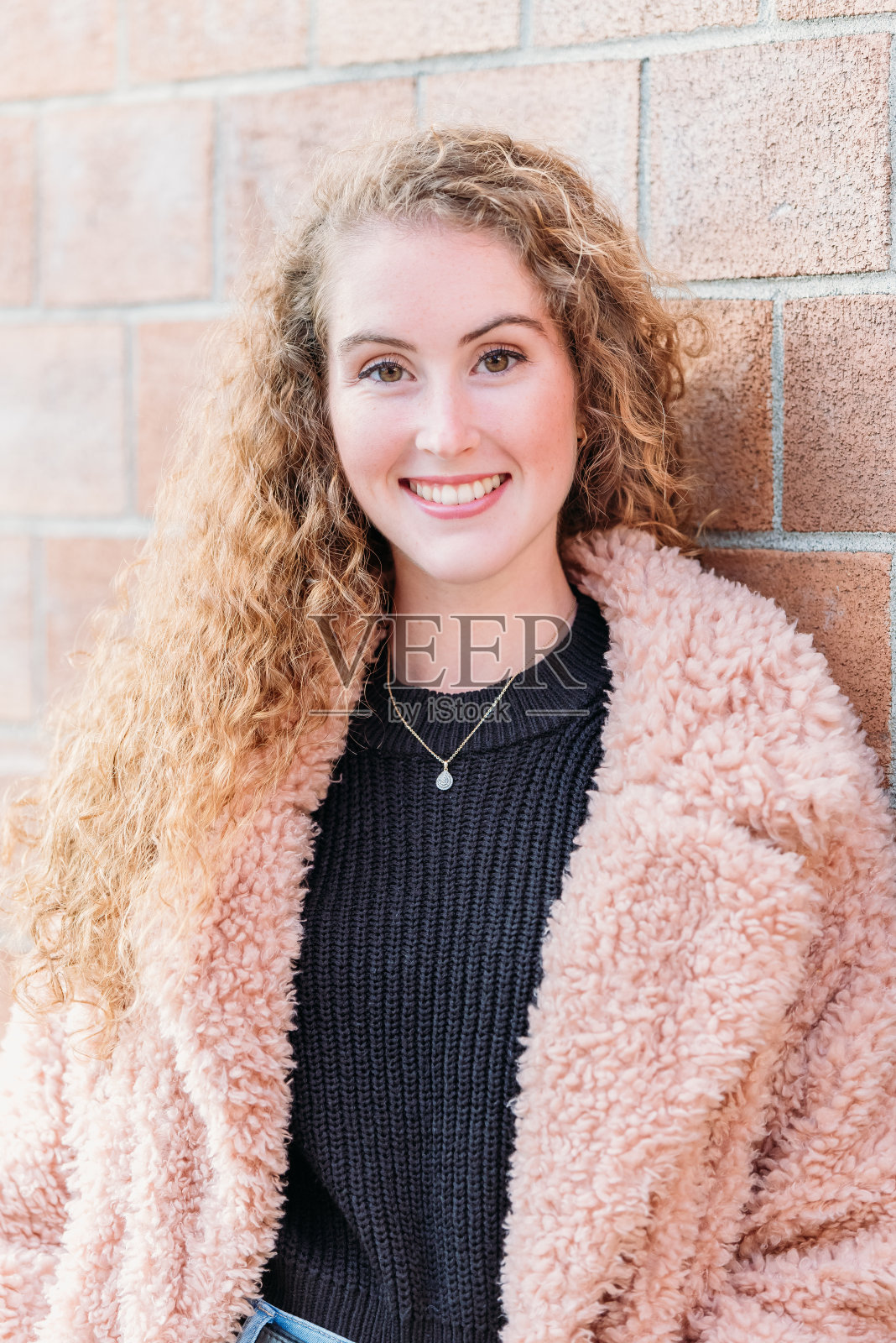 一位快乐的年轻女子倚在红墙上，穿着一件粉红色的毛衣。照片摄影图片