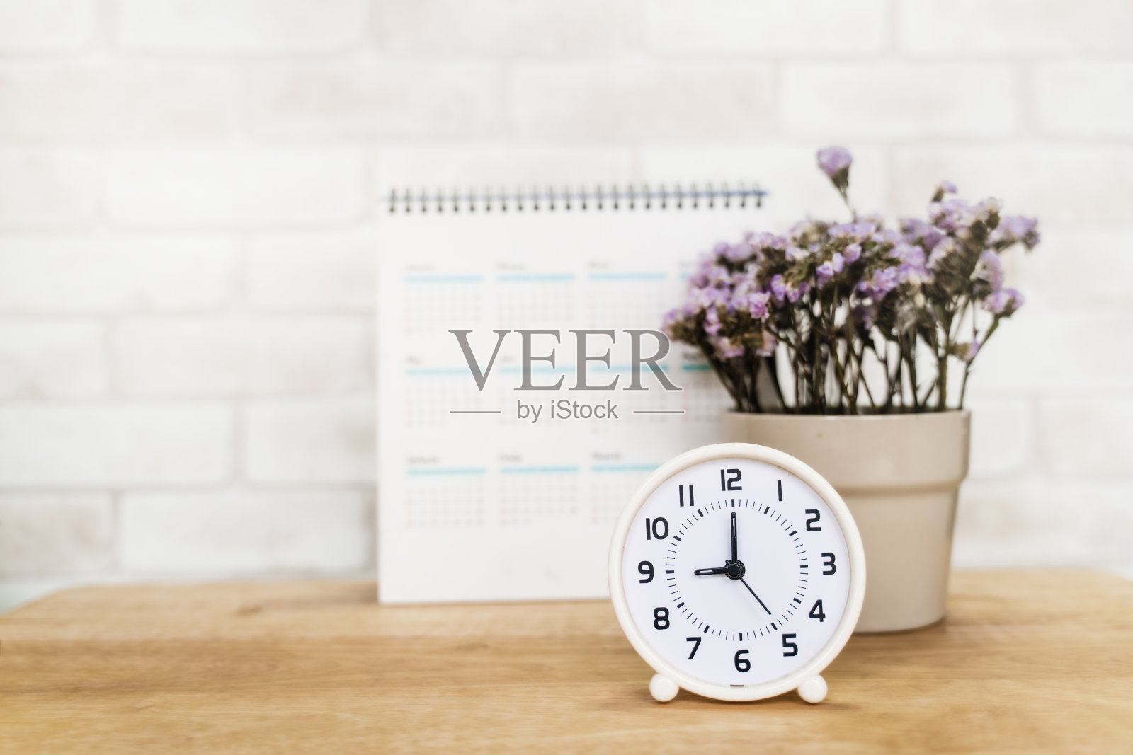 木制书桌上的陶瓷花瓶里放着白色的模拟时钟，上面有模糊的白色日历和紫色的花朵照片摄影图片