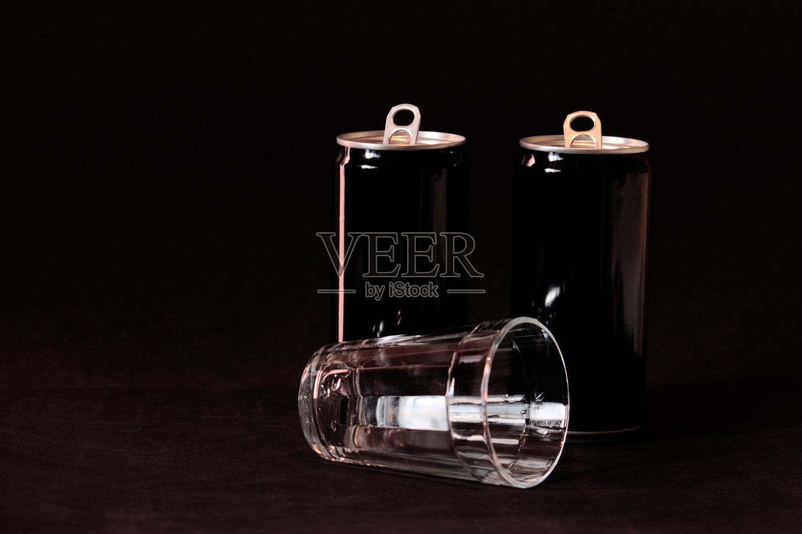 玻璃杯躺在两个涂成黑色的铝罐前面，在一个黑色的背景上。照片摄影图片