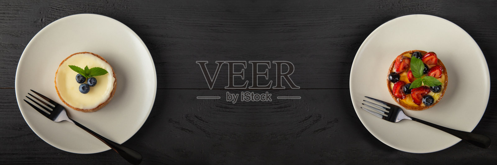 两个小挞或蛋糕，浆果，奶油和薄荷叶在白色的盘子和黑色的叉子，复制空间在黑色的木头，俯视图照片摄影图片