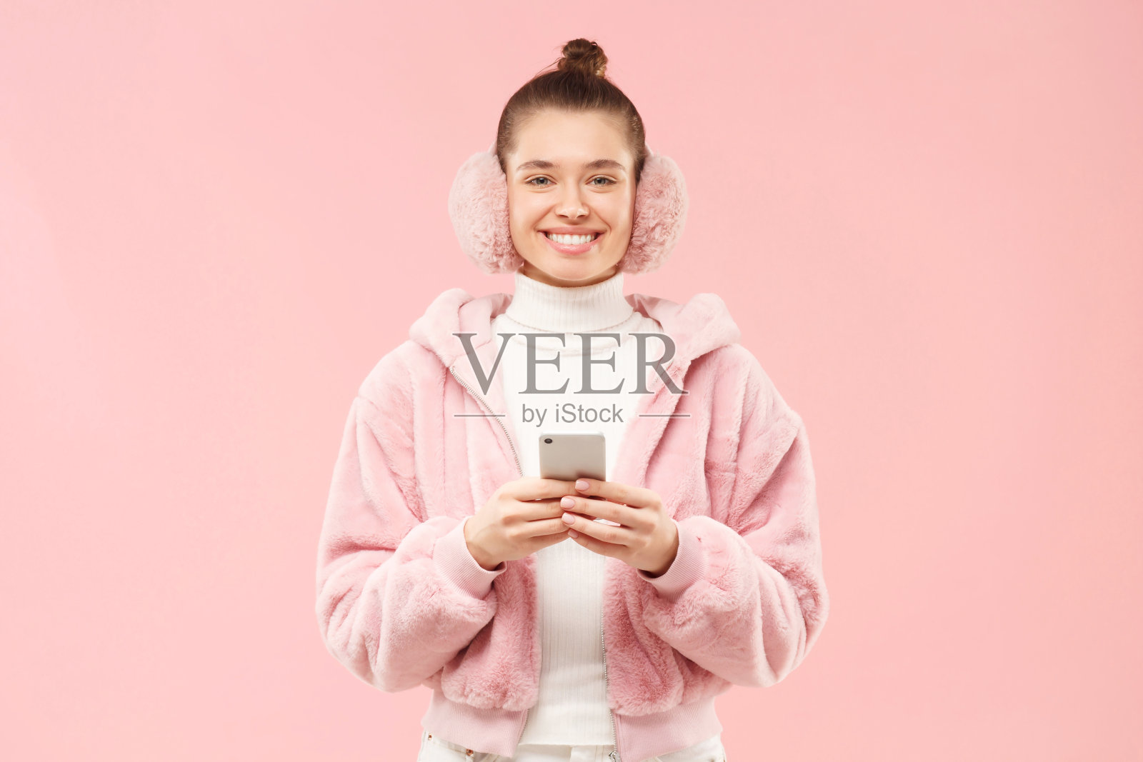 年轻女性微笑着，穿着冬天毛茸茸的外套，戴着耳套，手里拿着智能手机站着聊天或使用应用程序，在粉色背景下独处照片摄影图片
