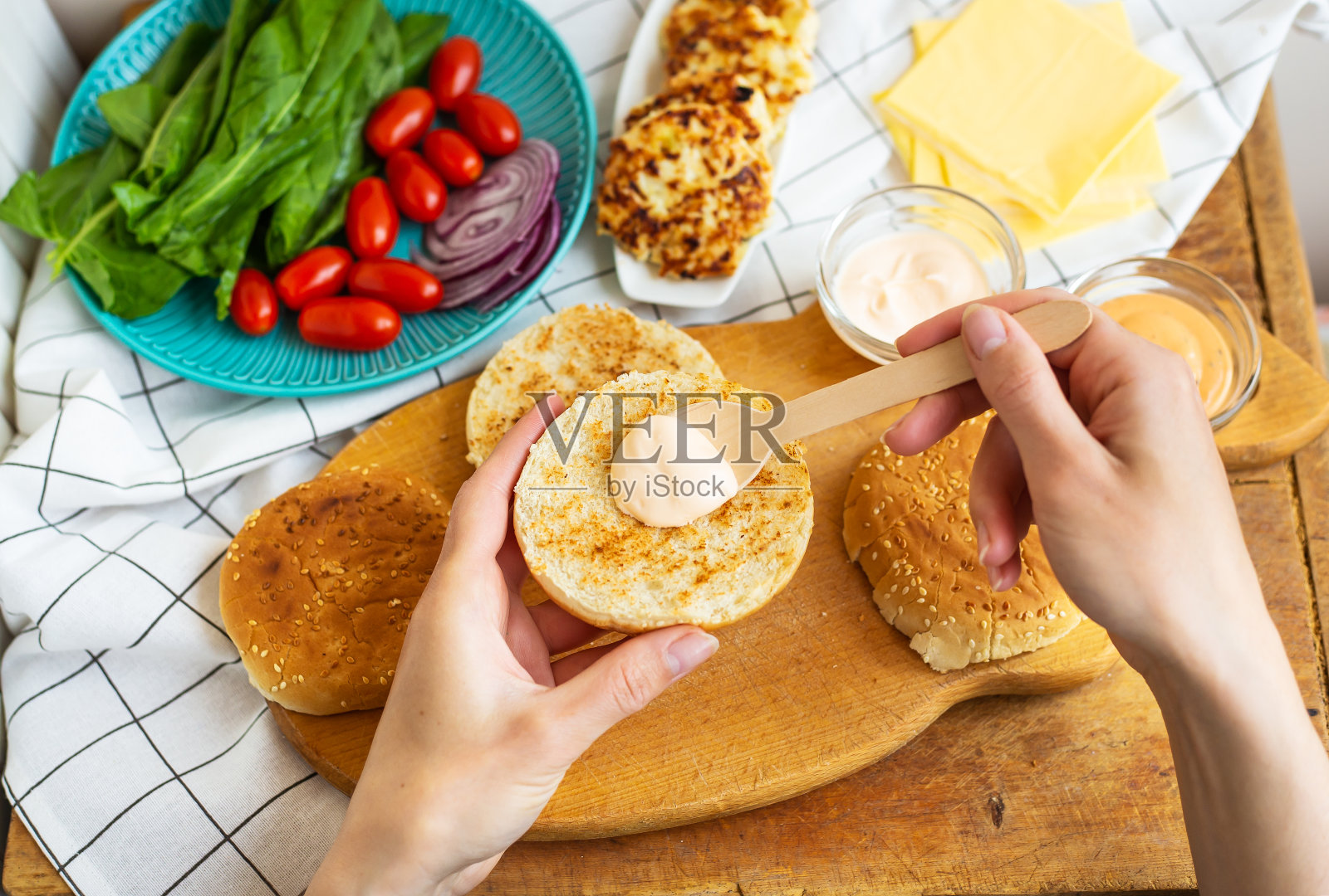 准备制作汉堡所需的所有材料-小圆面包，肉排，奶酪，沙拉，番茄，酱汁。俯视图，女孩的手拿着一个汉堡卷，并将酱汁洒在底部。照片摄影图片