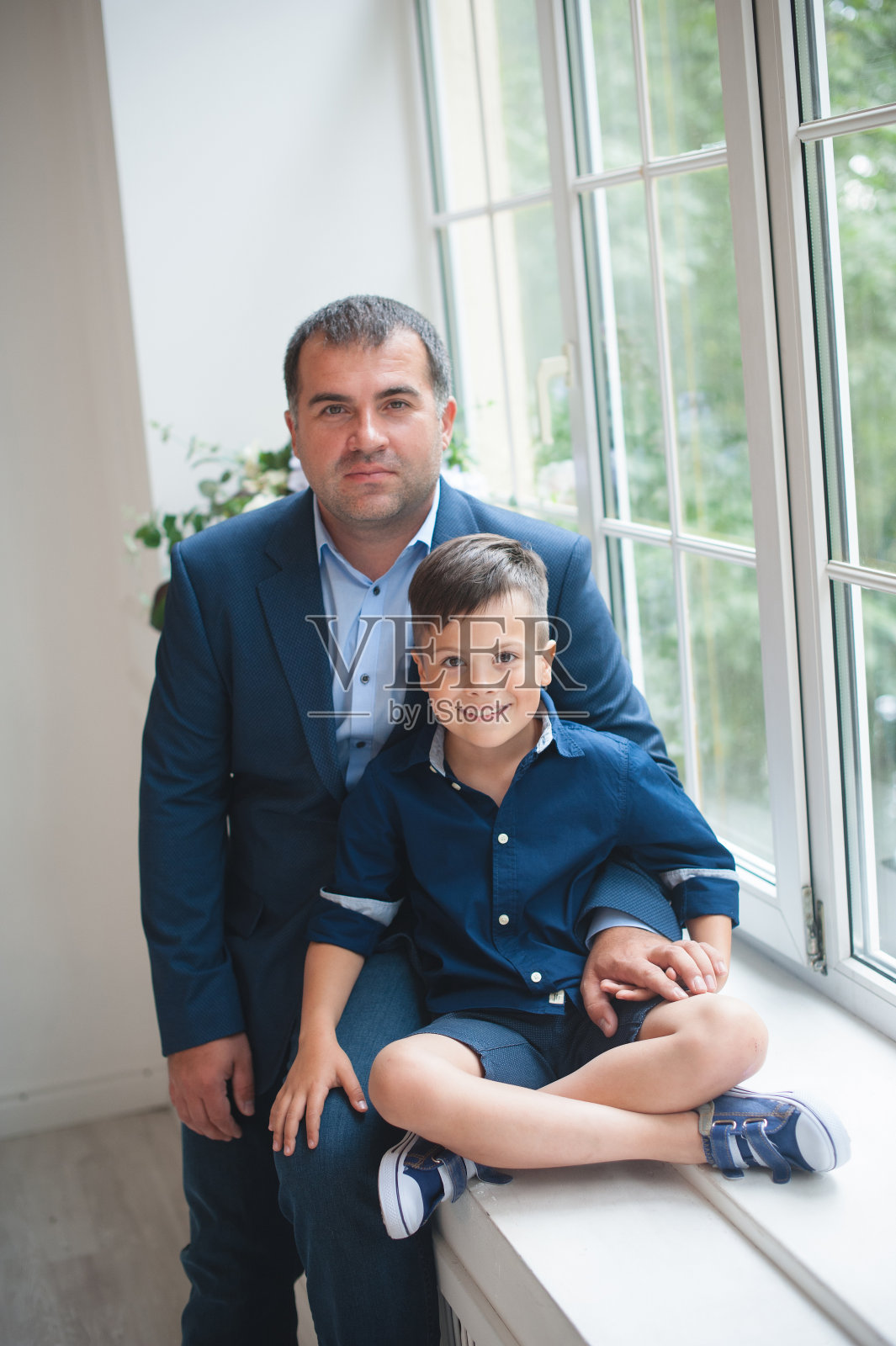 一个穿蓝色西装的男人和他的儿子坐在窗台上。庄严的事件,面带微笑。美丽的和愉快的。垂直图照片摄影图片