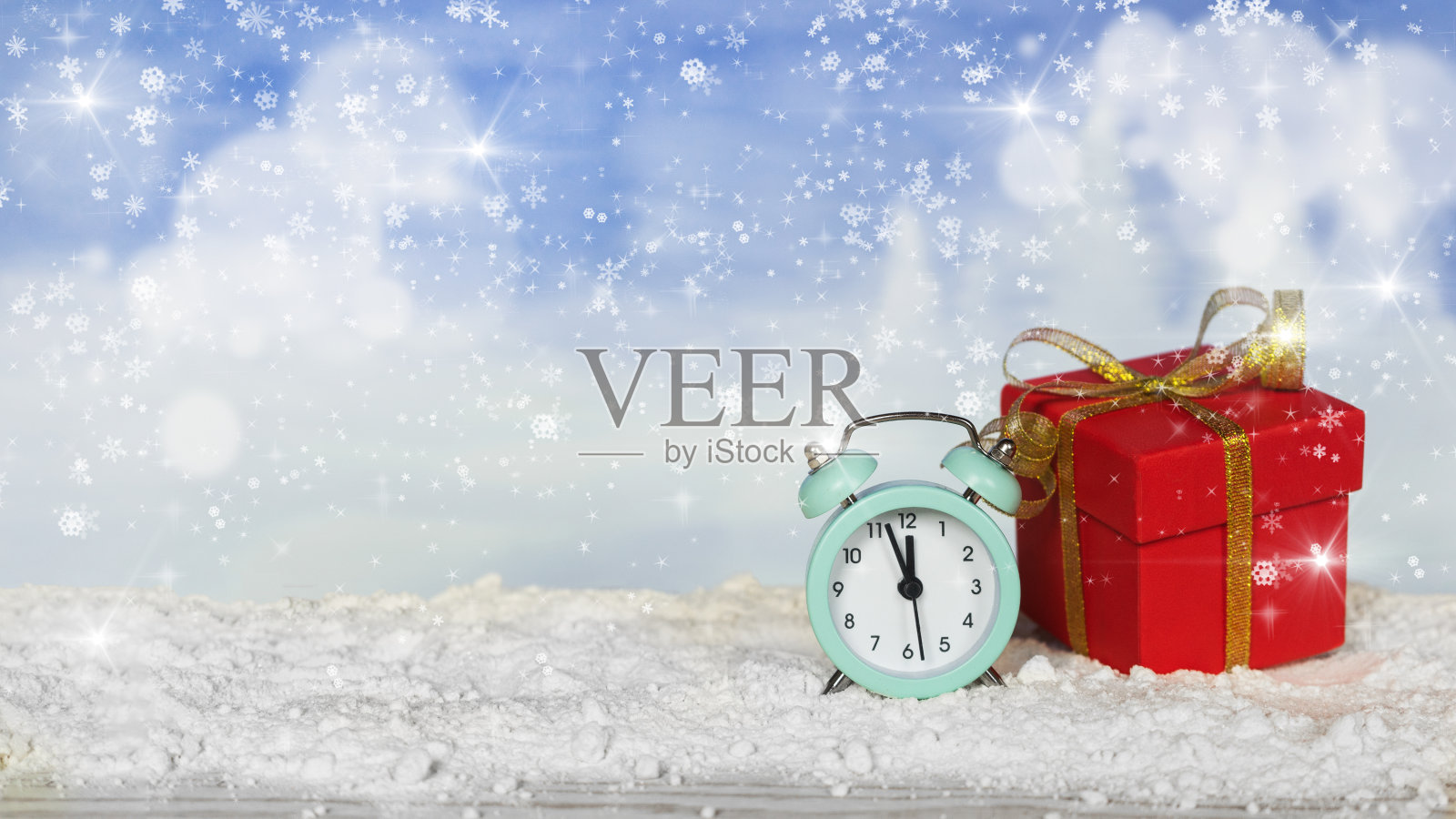 喜庆的圣诞标语。一个闹钟和一个带金色丝带的红色礼盒放在雪地上，背景是模糊的蓝色雪花。照片摄影图片