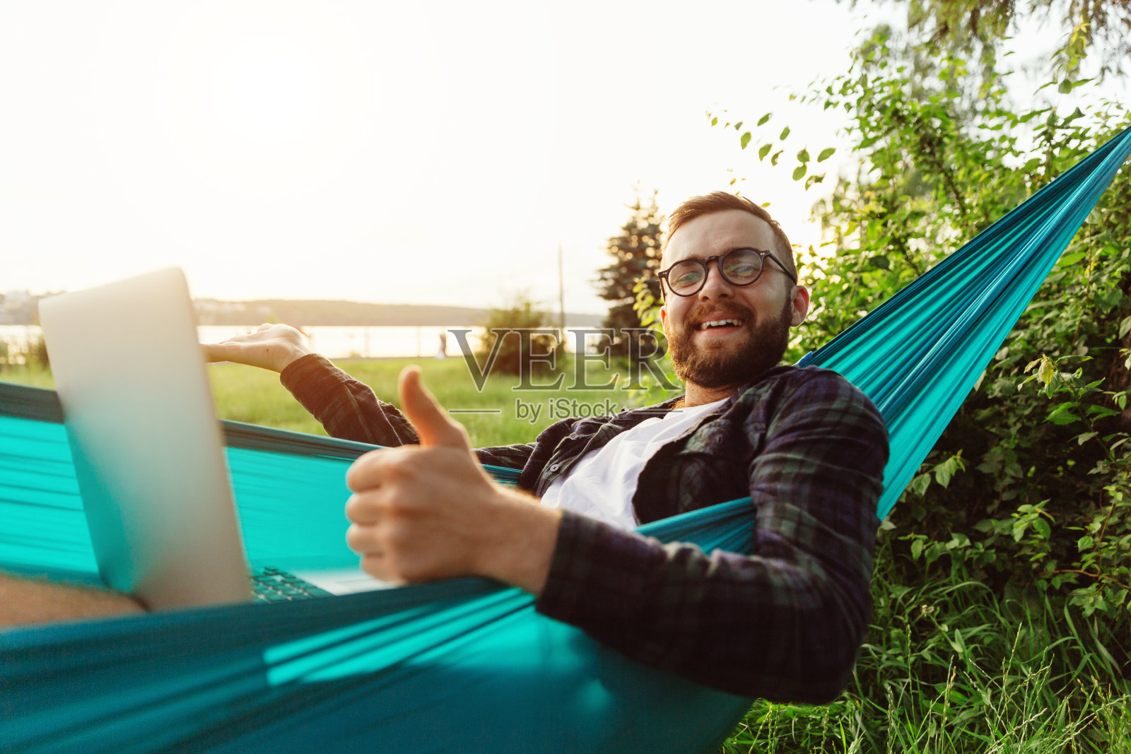 一名留着胡子、面带微笑的年轻男子躺在公园的吊床上，对着镜头满意地看着自己理想的工作照片摄影图片