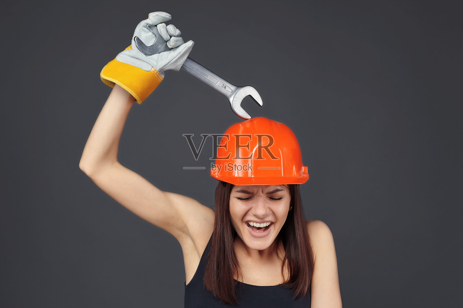 戴着橙色安全帽的建筑女孩用扳手砸到了头盔。介绍了防止机械损伤的概念。还有愤怒和非标准情绪压力的概念照片摄影图片
