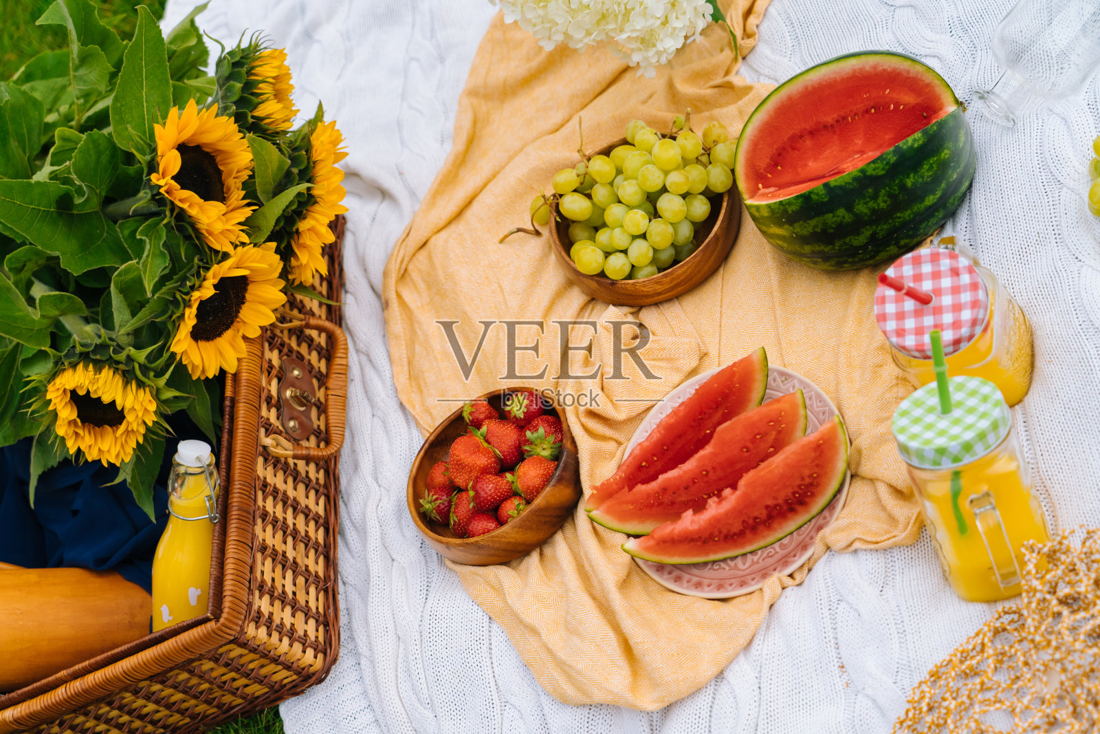 夏日野餐的概念在阳光明媚的日子有西瓜，水果，绣球花和向日葵花。草地上的野餐篮子，上面放着食物和提神的夏日饮料，上面盖着白色的针织毯子。有选择性的重点照片摄影图片