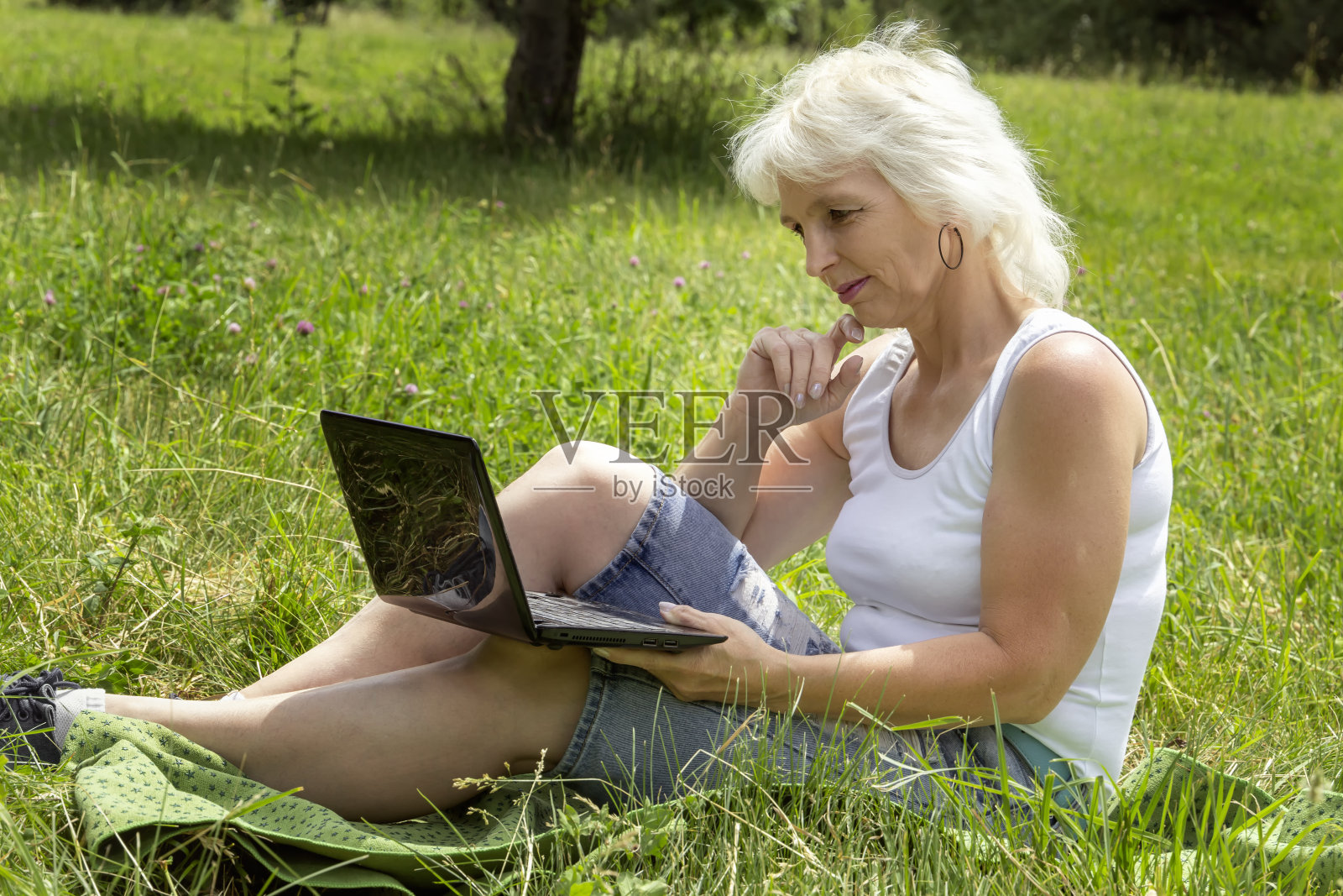 一位白发苍苍的苗条老人坐在公园阳光明媚的草坪上，通过笔记本电脑交流照片摄影图片