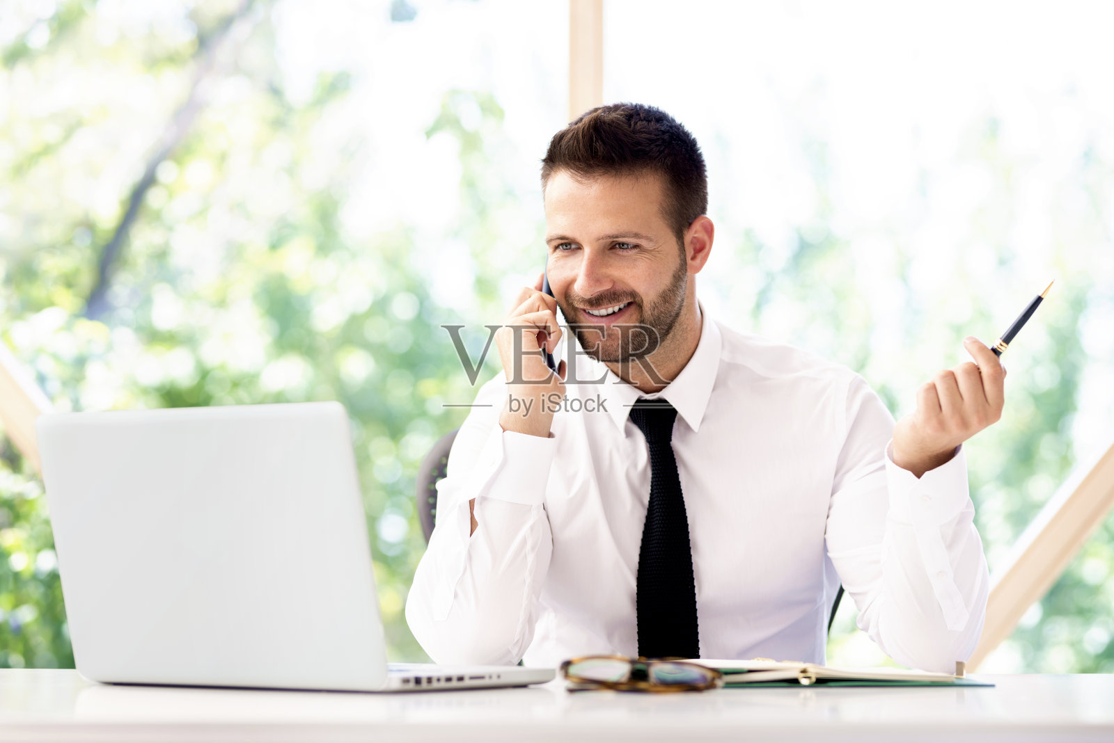 自信的商人坐在办公桌旁用手机和笔记本电脑与人交谈照片摄影图片