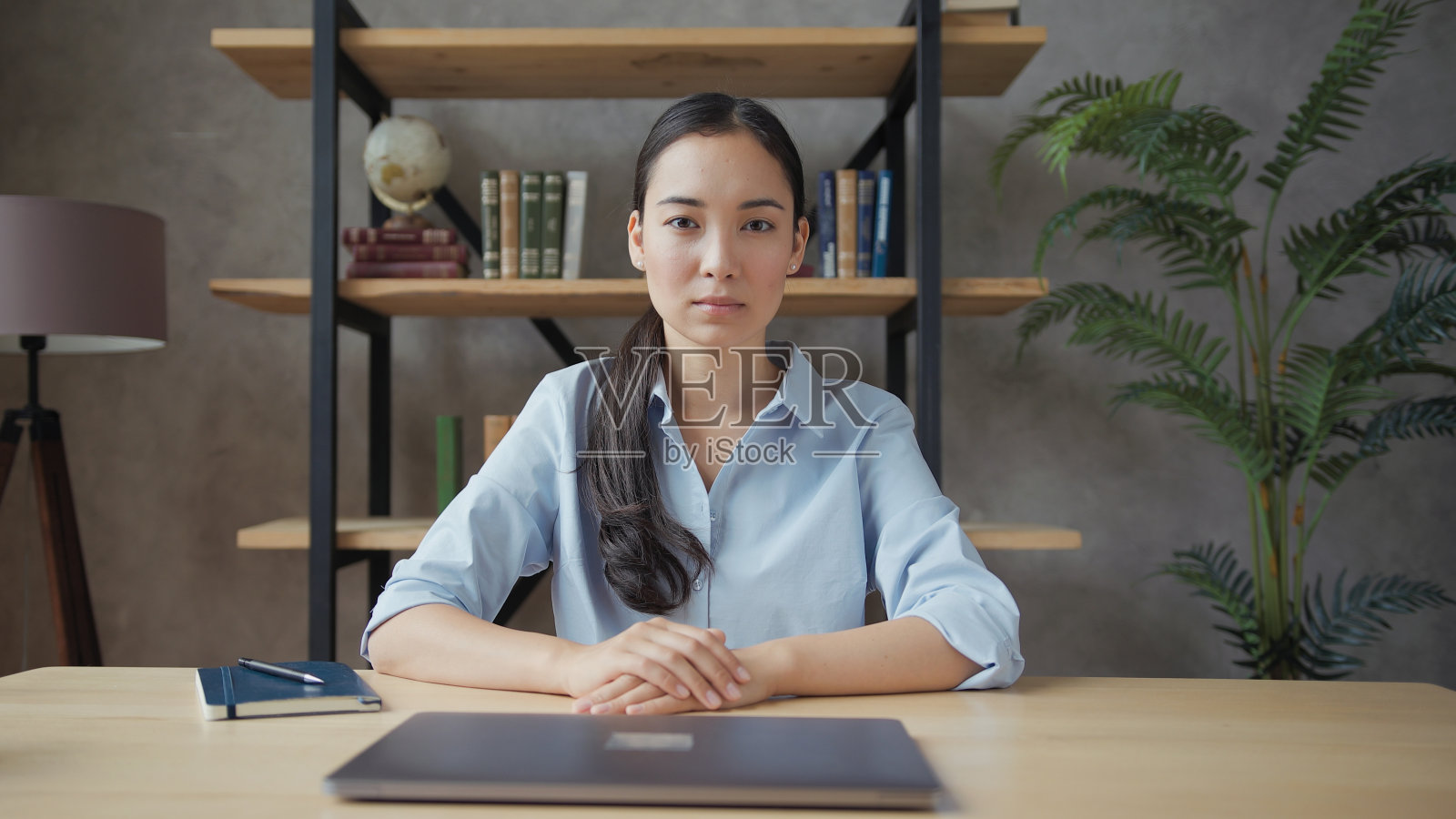 严肃的年轻亚洲女教师坐在桌子上，看着摄像机照片摄影图片