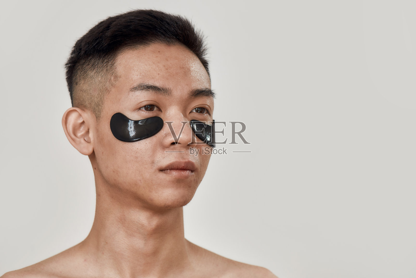 没有黑眼圈。赤裸上身的年轻亚洲男子的肖像与黑色的补丁下的眼睛看远孤立在白色的背景。美容，日常护肤，男士美容照片摄影图片