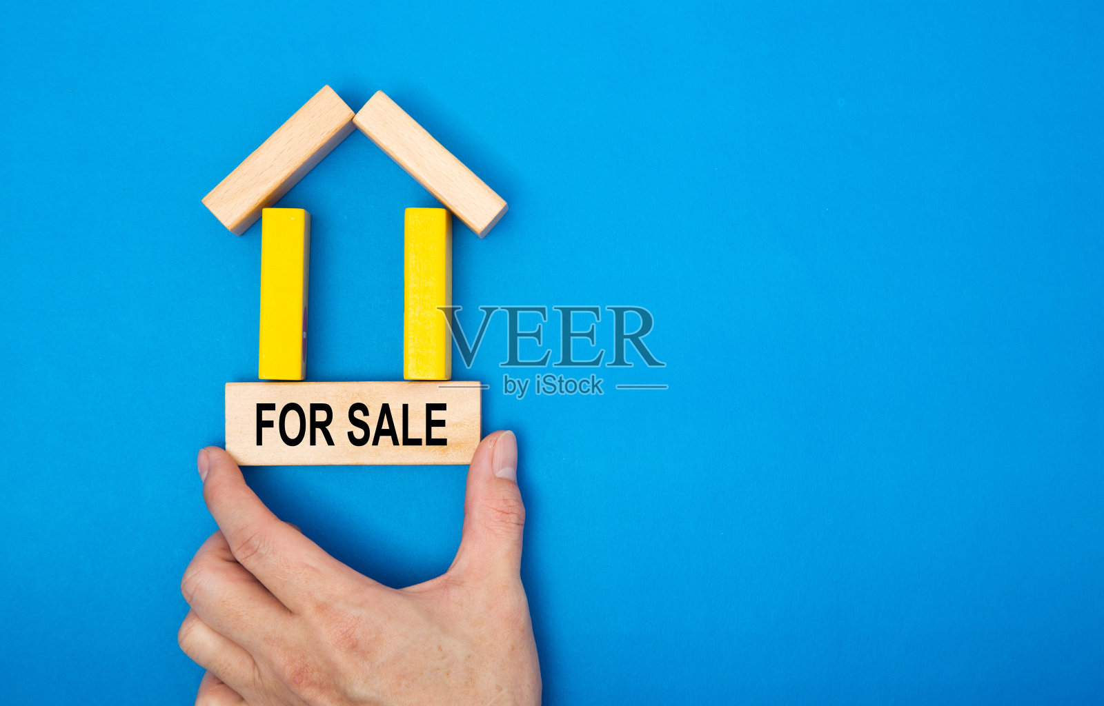 在房地产市场的概念形象中，一只手在一个用木块制成的房子里放置一个买卖标志。在蓝色背景。照片摄影图片