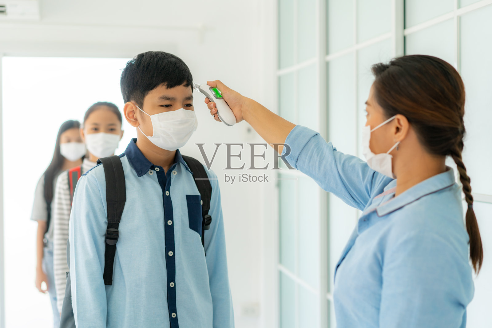 亚洲女教师在学生返校时使用体温计检查学生体温，以防止COVID-19的传播，新常态和教育理念。照片摄影图片