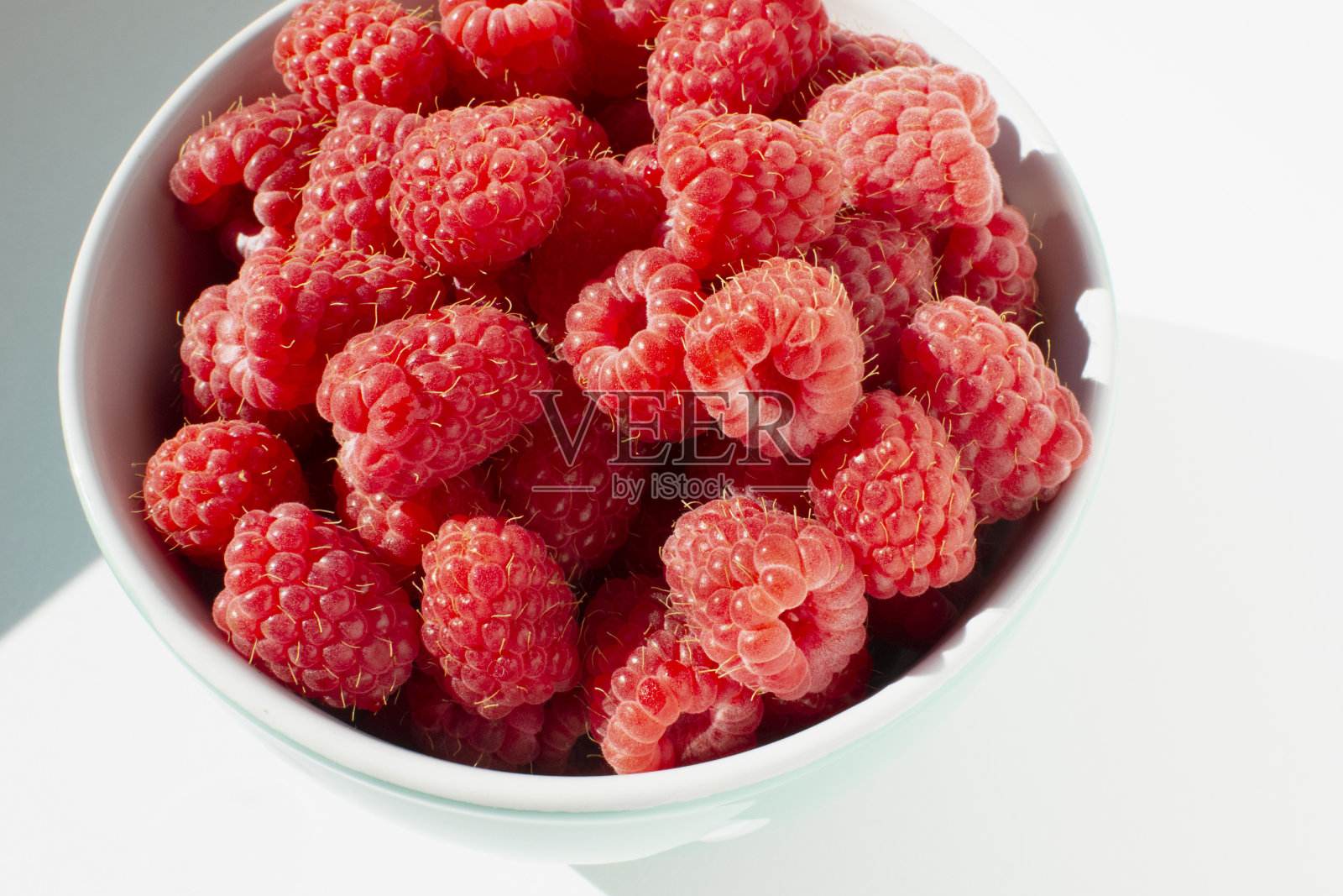 新鲜的覆盆子放在瓷碗里，放在白色的桌子上。特写照片与阳光。照片摄影图片