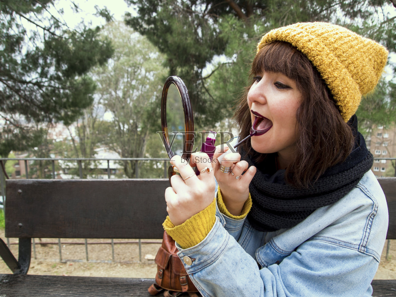 年轻的成年女子，戴着黄色的羊毛帽，坐在公园的长椅上，对着镜子画嘴唇照片摄影图片
