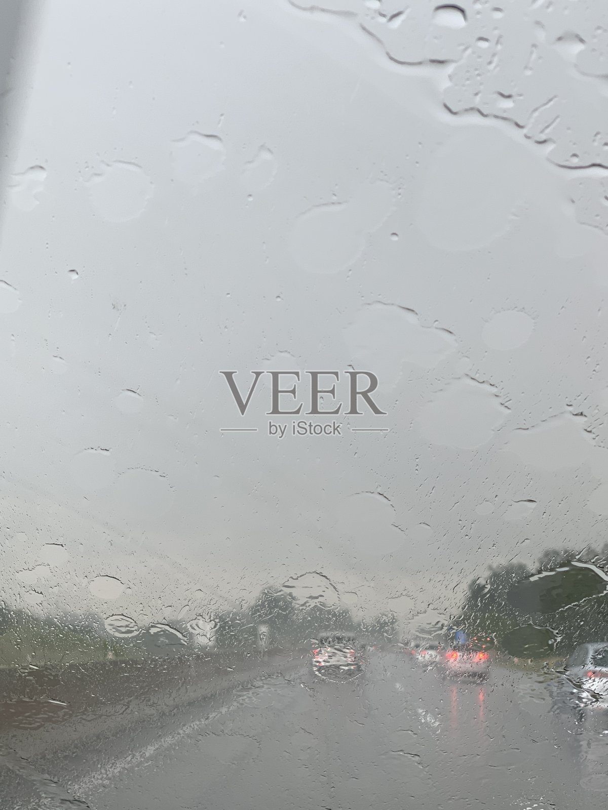 在大雨中，高速公路上一辆汽车的挡风玻璃上覆盖着一层薄薄的水膜。周围的风景是模糊的，有适当照明的汽车在前面行驶。照片摄影图片