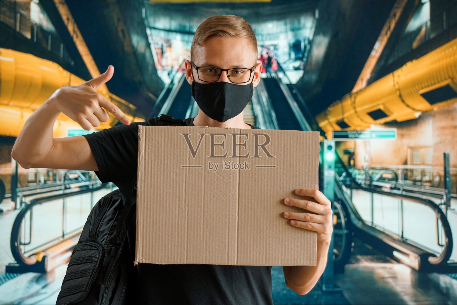 一名戴着黑色医用口罩、背着背包、手里拿着空牌子的年轻男子站在机场的自动扶梯附近照片摄影图片