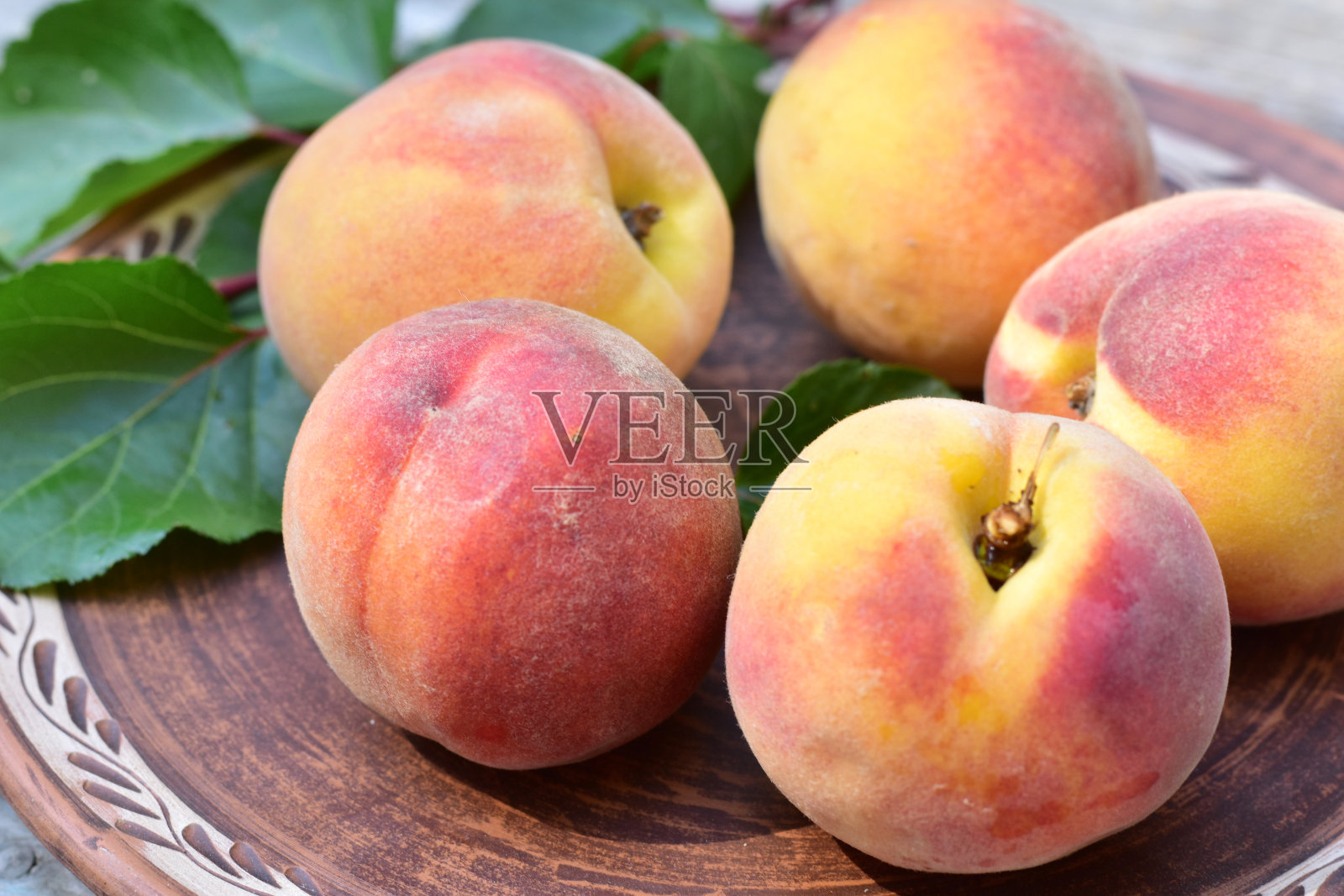 粉红色的、多汁的、成熟的桃子躺在一个粘土盘子里，夹在绿叶中间。照片摄影图片