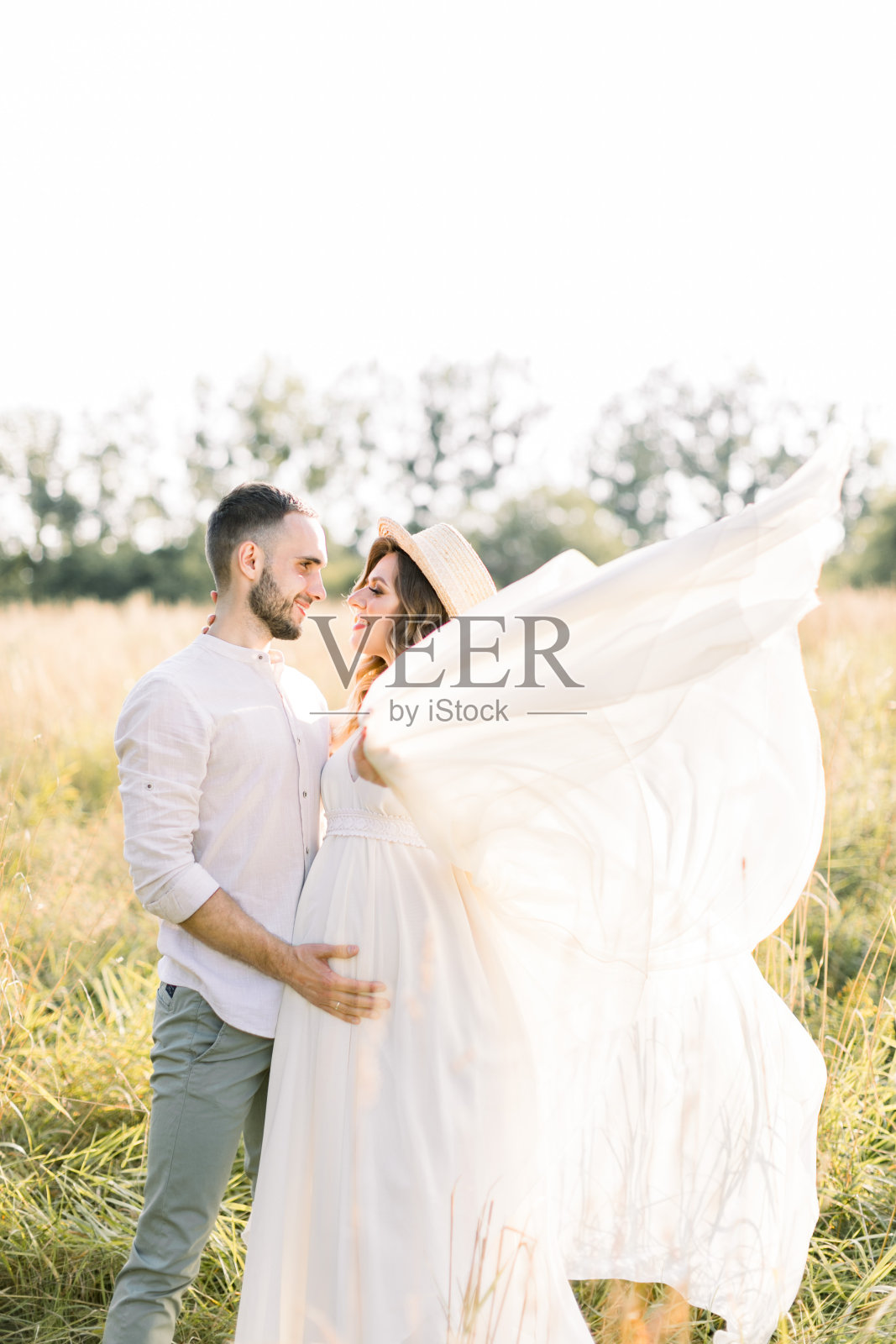 英俊的络腮胡男人和他漂亮的怀孕妻子在日落时分在户外田野里散步。浪漫家庭，怀孕概念照片摄影图片