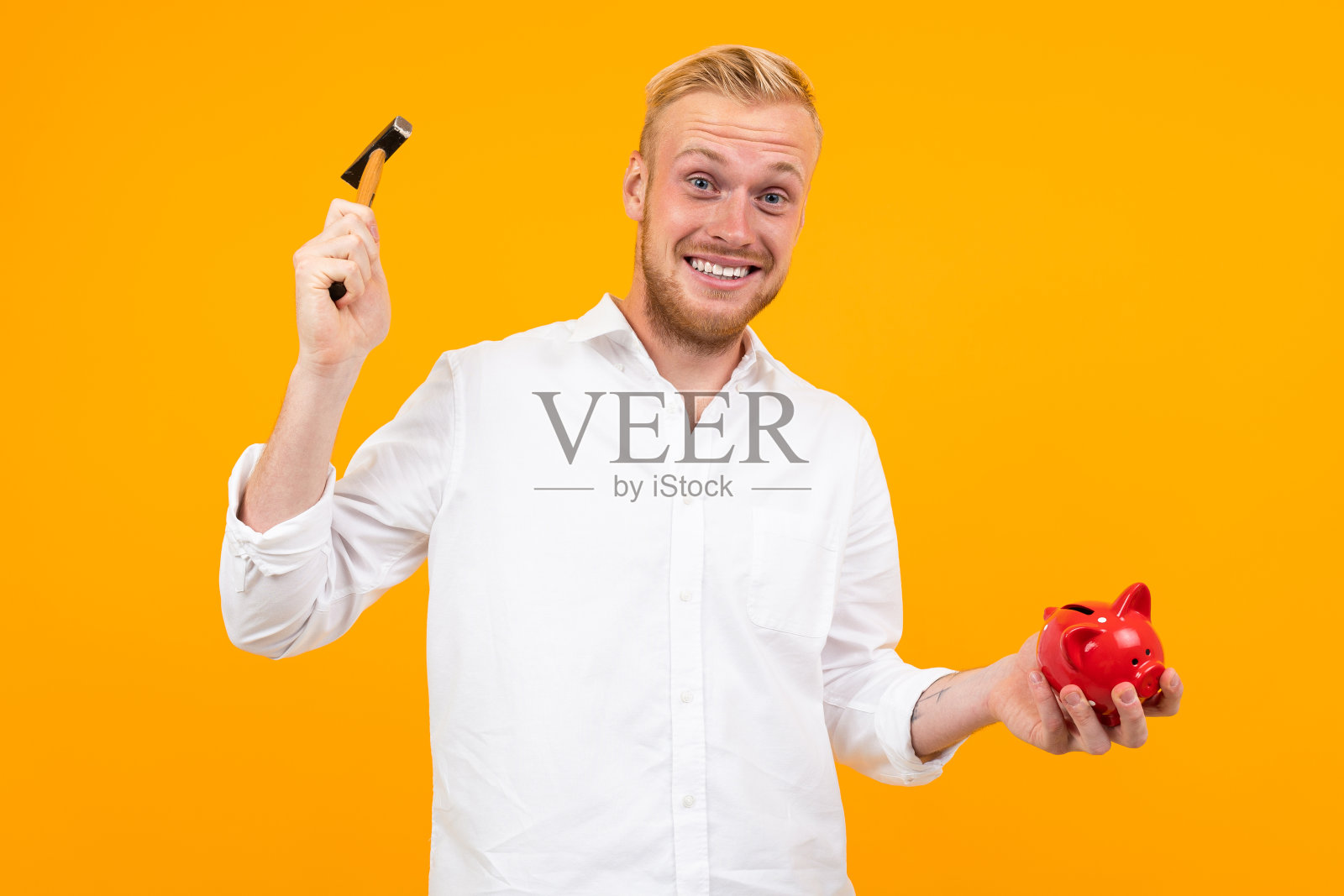 一个穿白衬衫的金发男子用一把锤子在黄色背景上砸存钱罐。照片摄影图片