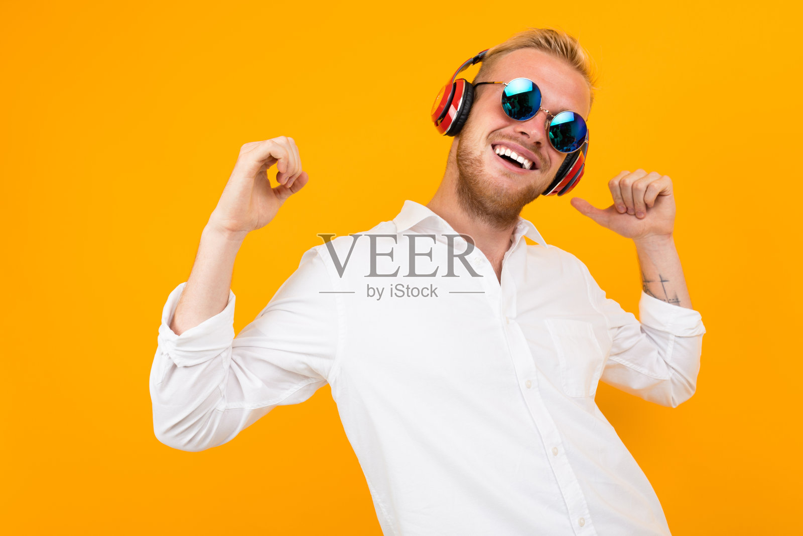 欧洲男人穿着白色衬衫，戴着太阳镜，戴着黄色背景的大耳机听音乐。照片摄影图片