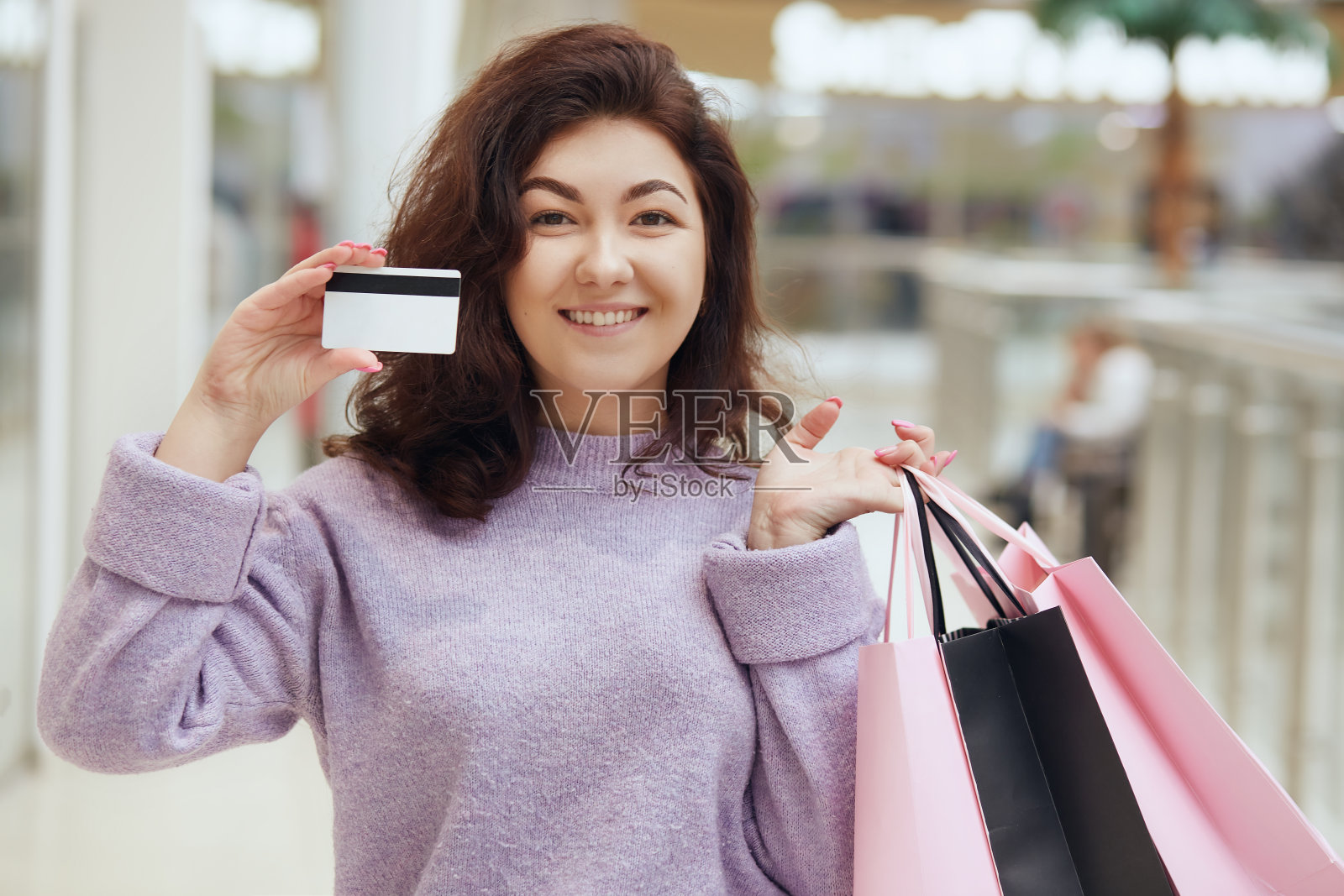 年轻迷人的女性与黑色波浪发摆姿势在购物中心，手里拿着购物袋和信用卡，支付她的购买与她的信用卡。照片摄影图片