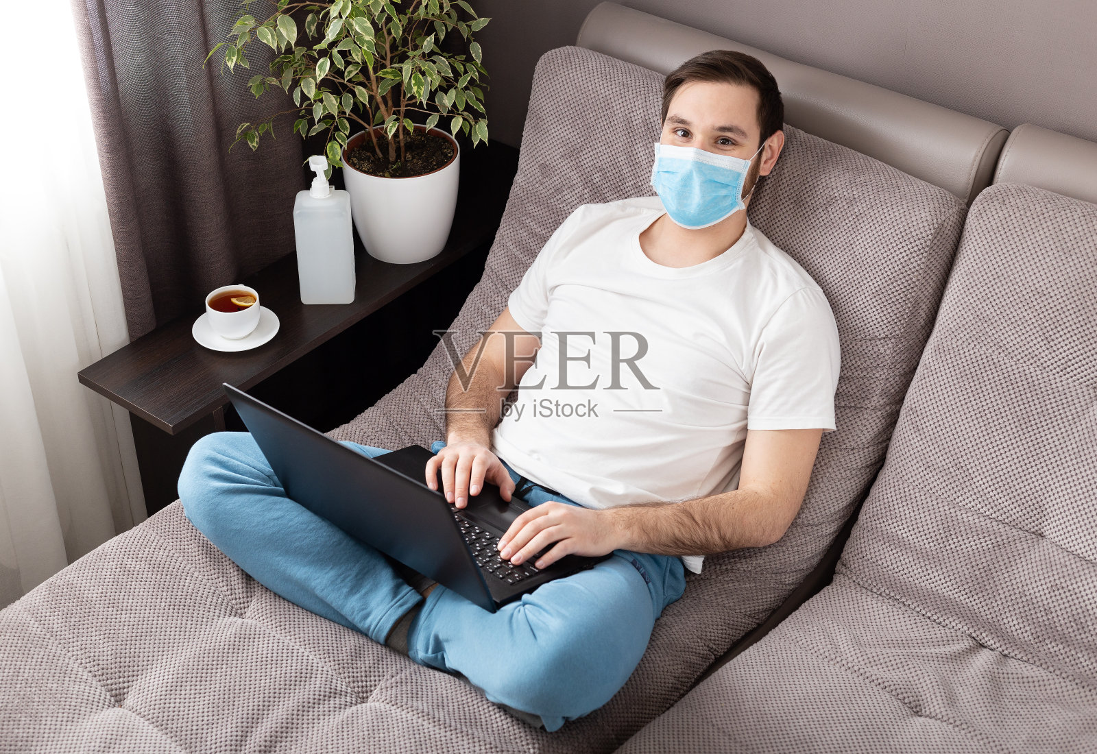快乐放松的年轻白人男子在家工作戴着防护口罩使用笔记本电脑。舒适的家庭办公室，在冠状病毒大流行期间在沙发上工作，冠状病毒19隔离。远程工作,自由职业者照片摄影图片