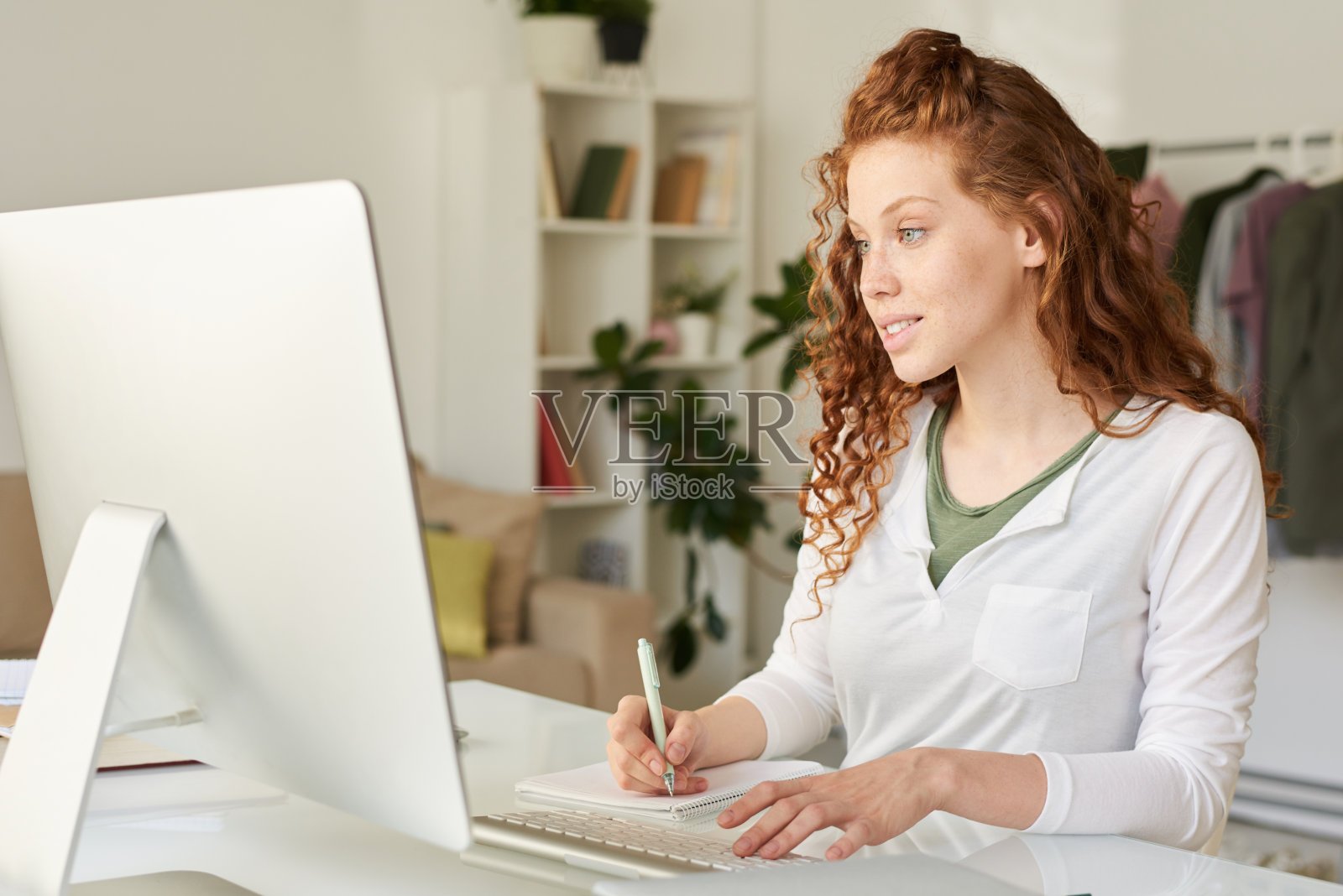 微笑迷人的红头发学生女孩坐在桌子上，做笔记，同时享受在线课程与专业的老师照片摄影图片