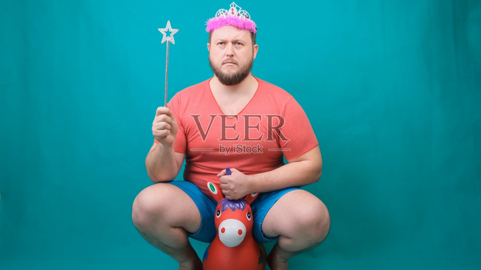 一名身穿粉色t恤、头戴耳坠、蓄着胡须的英俊古怪男子手持魔杖骑着独角兽。一个有趣的巫师笑话，用来实现一个愿望照片摄影图片