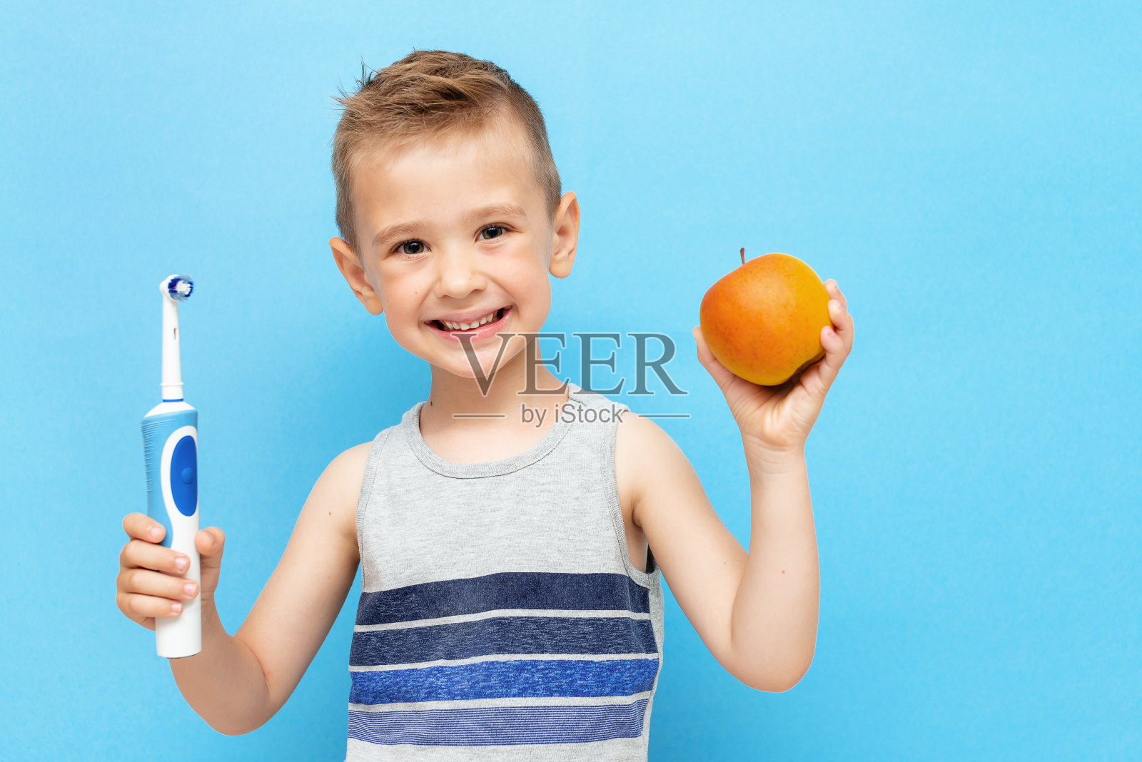 一个微笑的小男孩，手里拿着一把电动牙刷和一个苹果，在蓝色背景上照片摄影图片