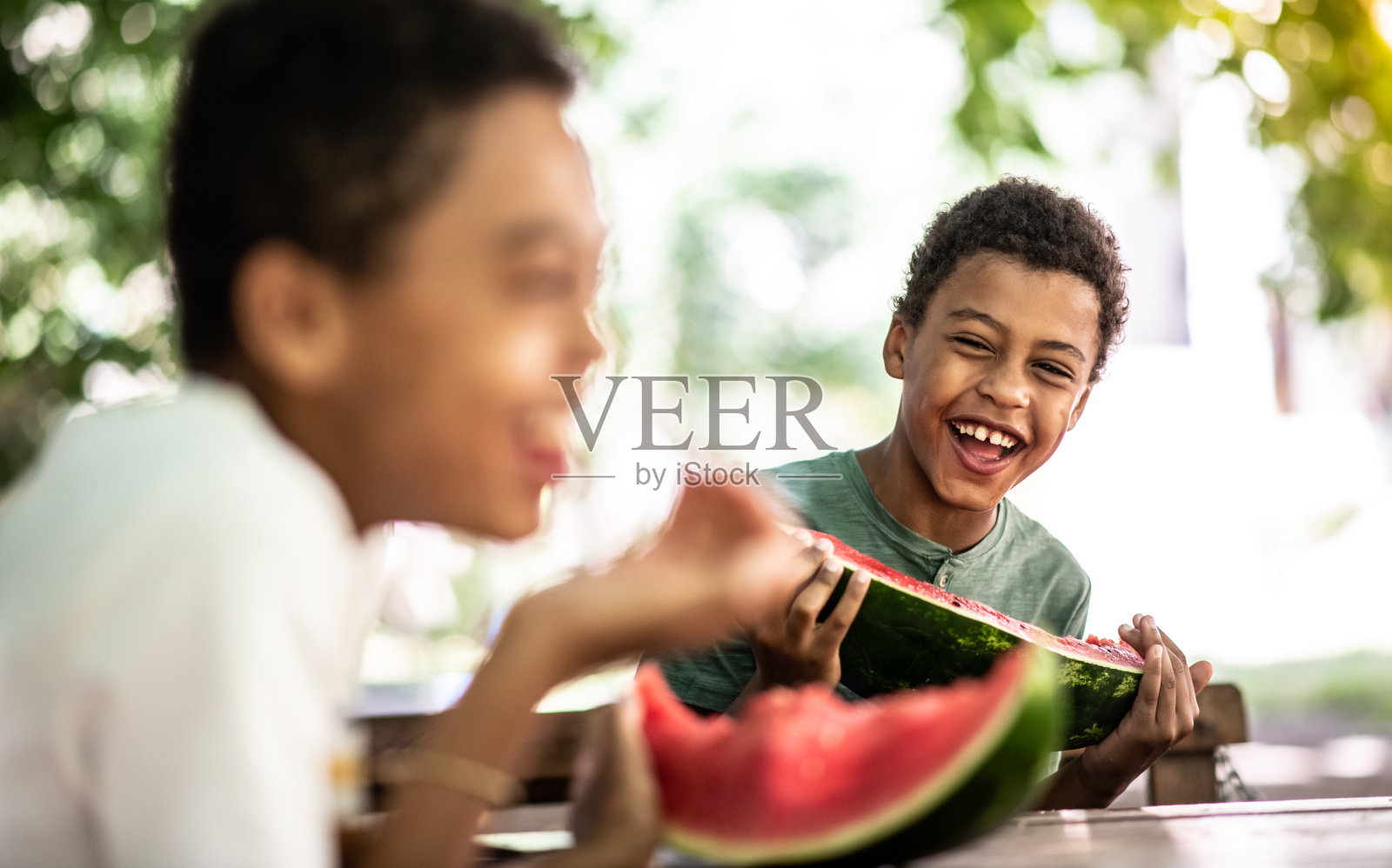 两个男孩坐在长椅上，在一个阳光明媚的日子里吃着西瓜，他们笑了照片摄影图片