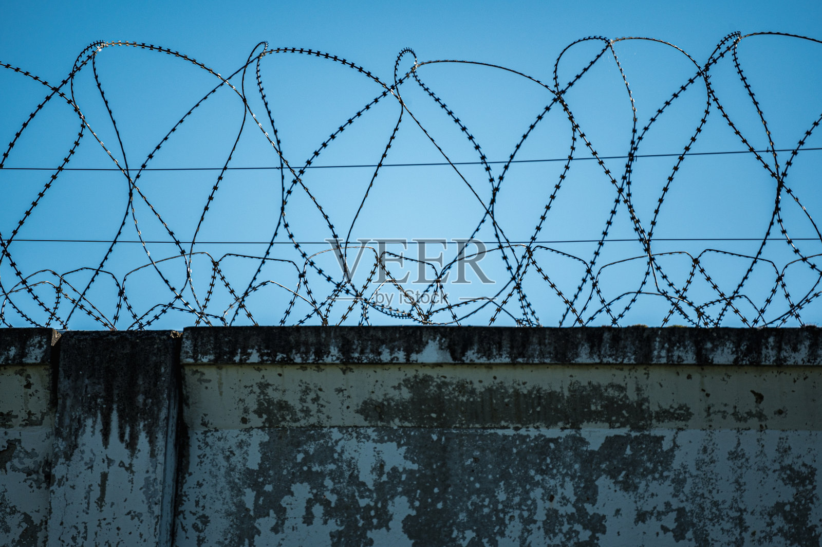 带带刺铁丝网的混凝土栅栏。蓝天上的监狱高墙上挂着带刺的铁丝网。照片摄影图片