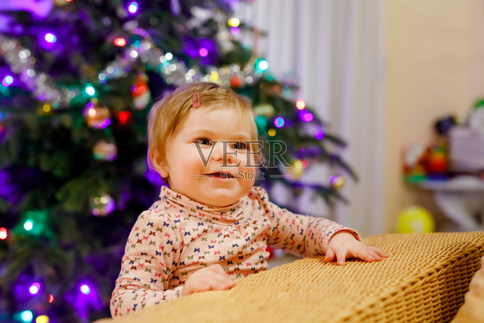 小可爱的小女孩学习走路和站立的肖像。背景是圣诞树和彩灯。可爱的蹒跚学步的女孩在家。穿着休闲装的漂亮宝宝照片摄影图片