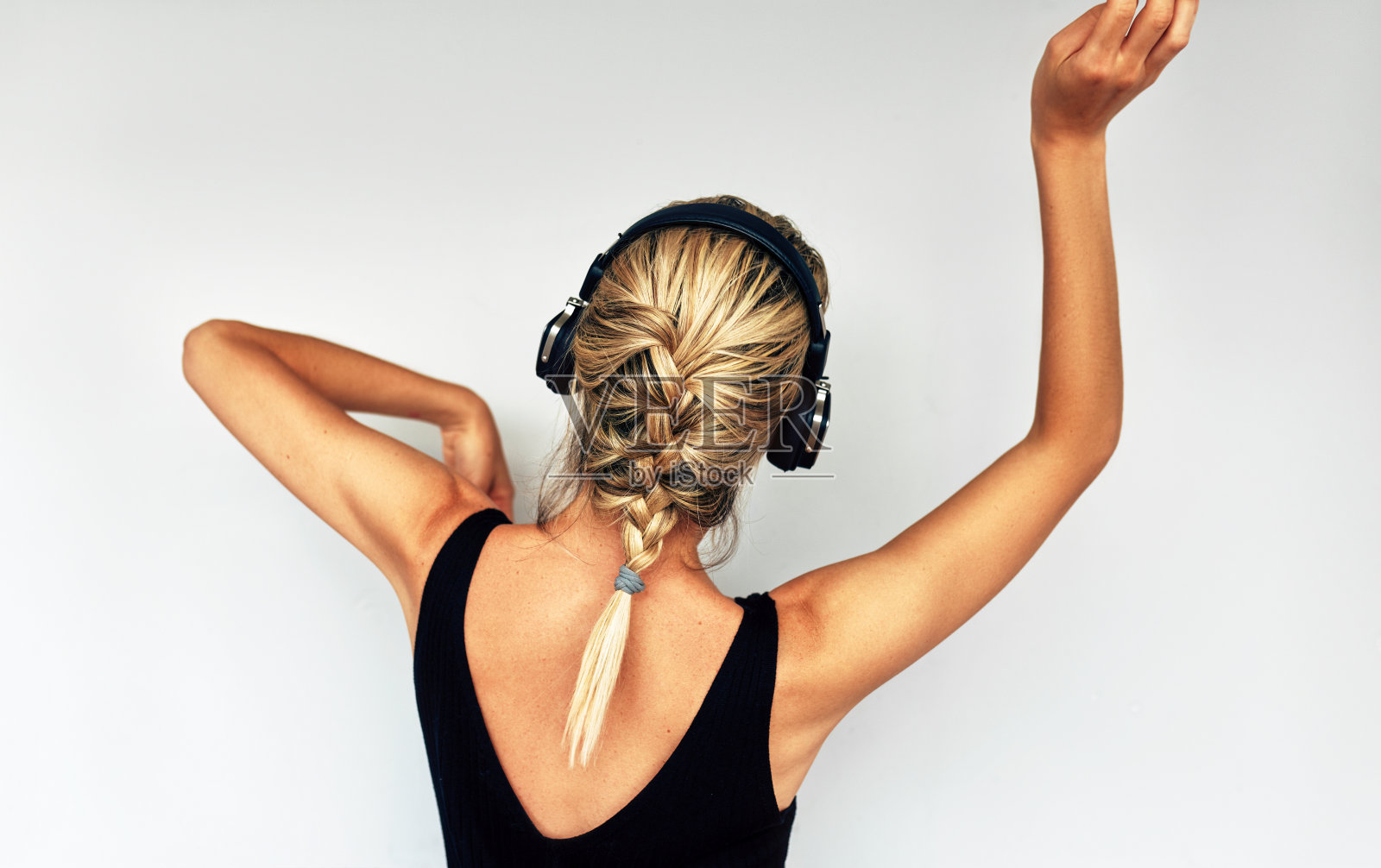 美丽的后视图有吸引力的金发年轻女子跳舞和听音乐耳机。快乐的女性穿着黑色裙子，梳着辫子，戴着耳机在录音室里听着最喜欢的歌曲照片摄影图片