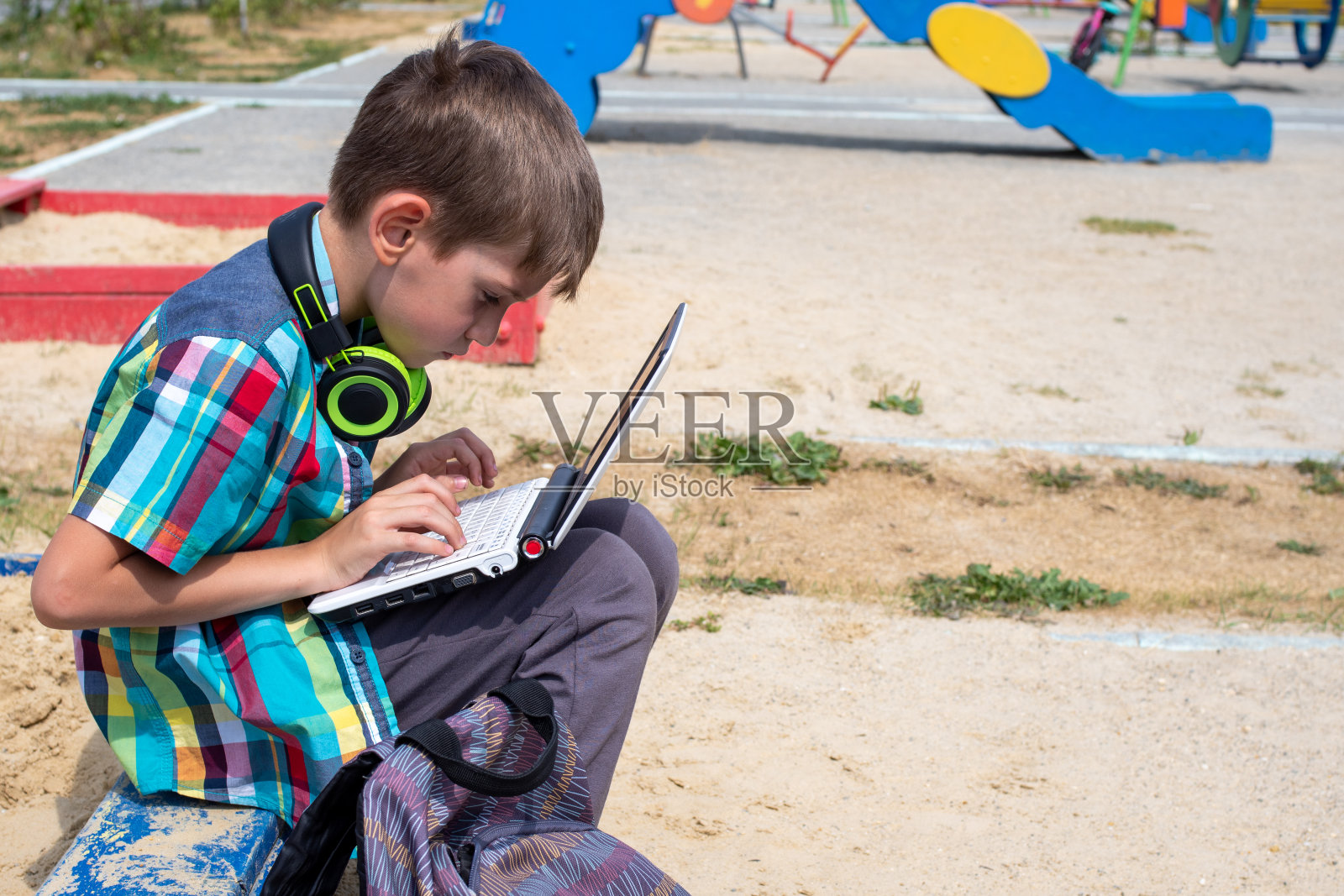 一个穿着休闲服的可爱男孩正在操场上玩笔记本电脑，旁边放着一个书包照片摄影图片
