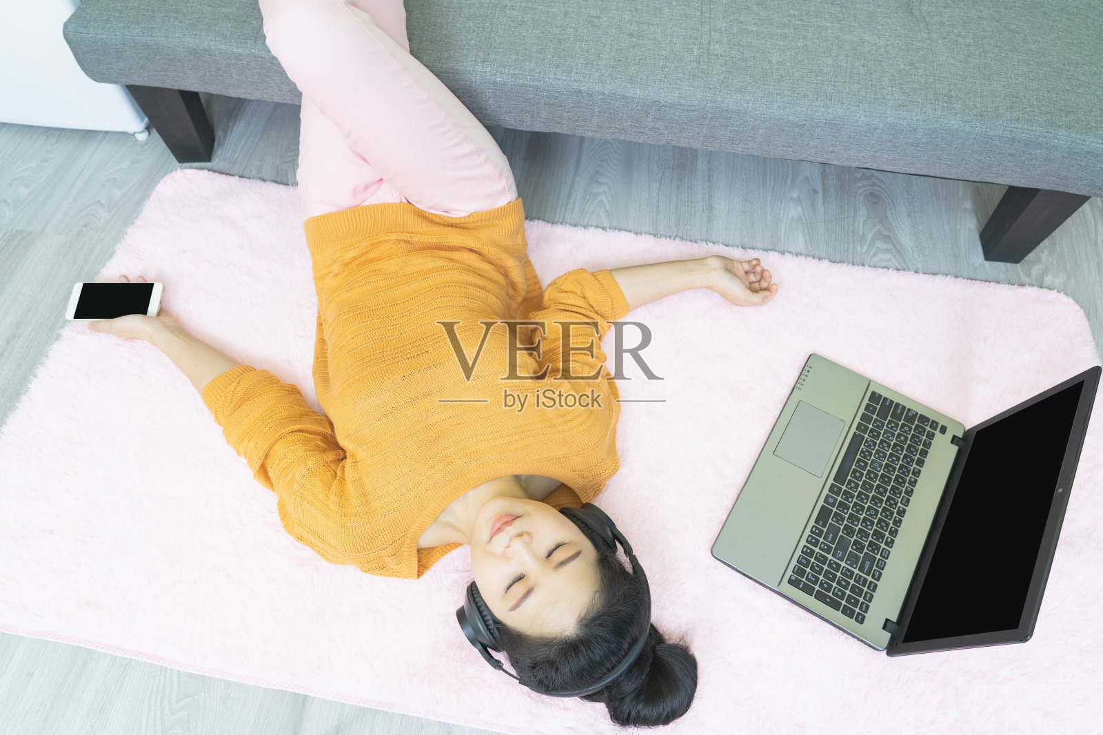 人与休闲概念——快乐的年轻亚洲女子戴着耳机躺在粉红色的地毯上，用笔记本电脑听音乐，在家里享受照片摄影图片