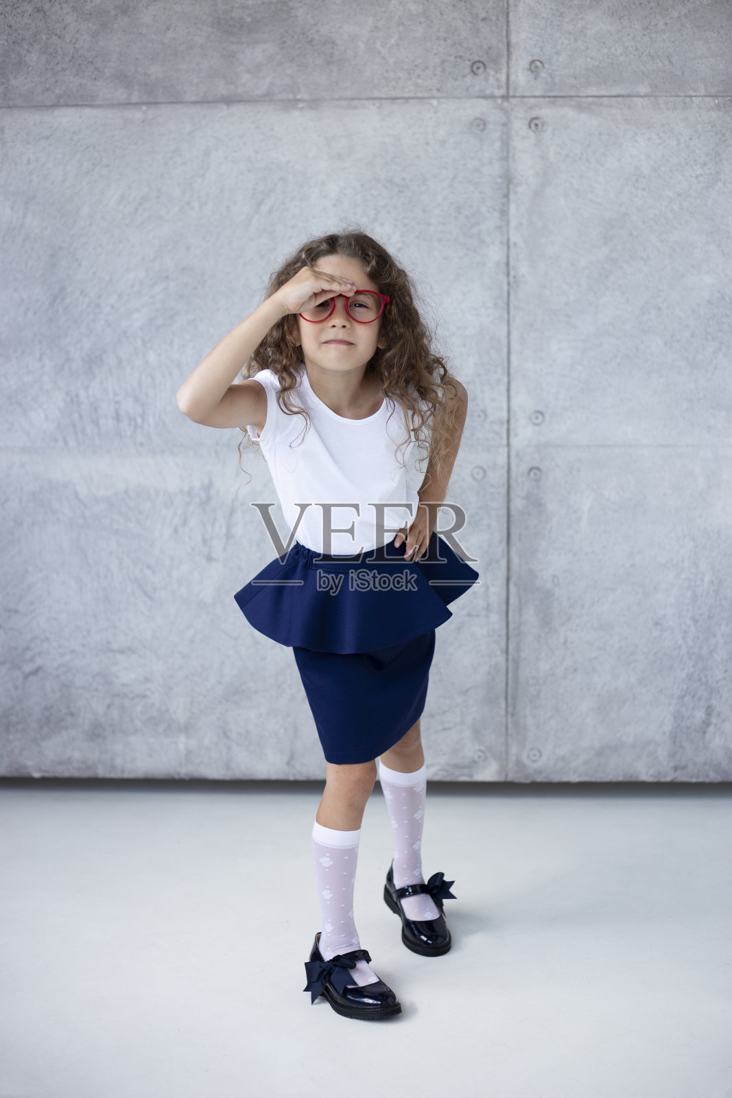 一个可爱的卷发小女孩，穿着裙子，戴着红色眼镜，在具体的背景上。照片摄影图片