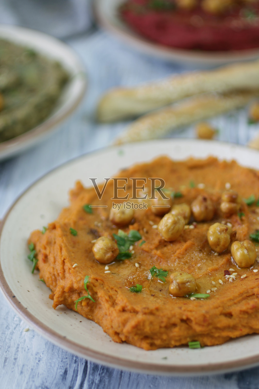 碗里有甜菜根、晒干的番茄和罗勒鹰嘴豆泥，周围是干烤鹰嘴豆和铺着芝麻的自制面包条，背景是白色的木头，希腊美食，重点放在前景照片摄影图片