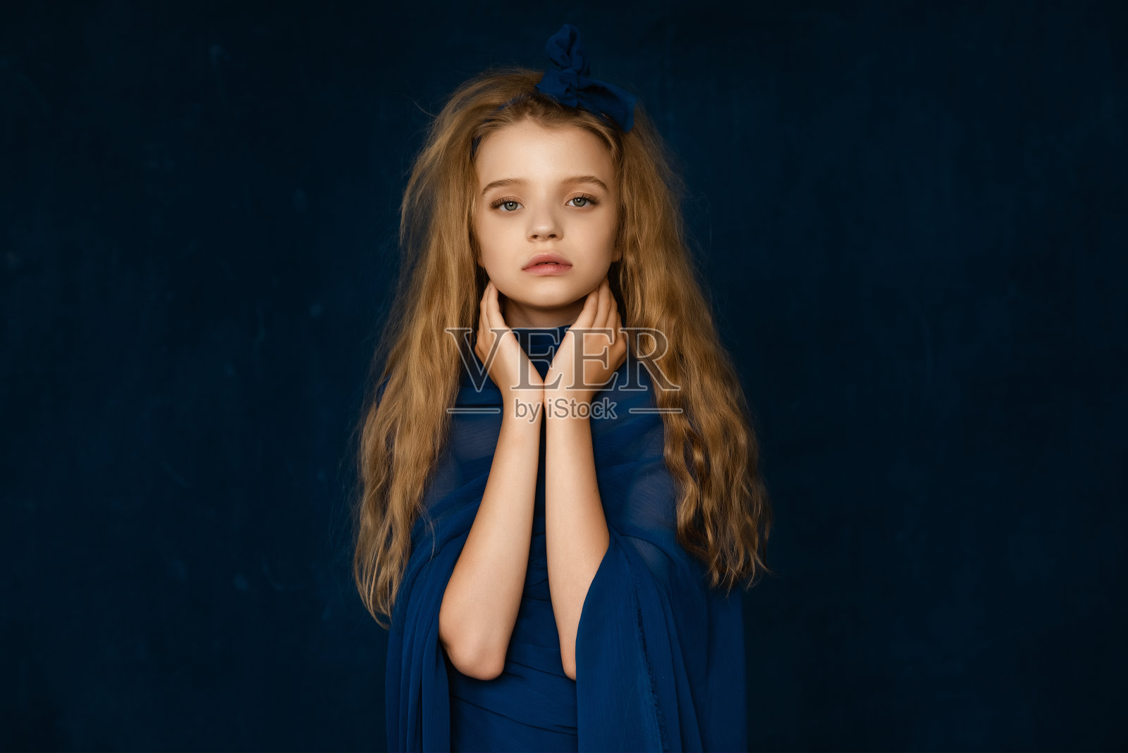 美丽的年轻女孩的艺术肖像10-12岁，金色卷曲的长发，在蓝色背景上她的手在她的脸上照片摄影图片