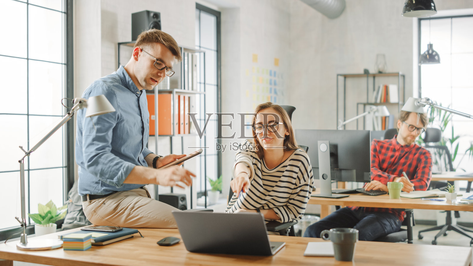 男同事强势地坐在一位年轻女同事面前的桌子上，手里拿着平板电脑，上面写着商业笔记。创意公司的友好氛围。他们讨论商业想法照片摄影图片