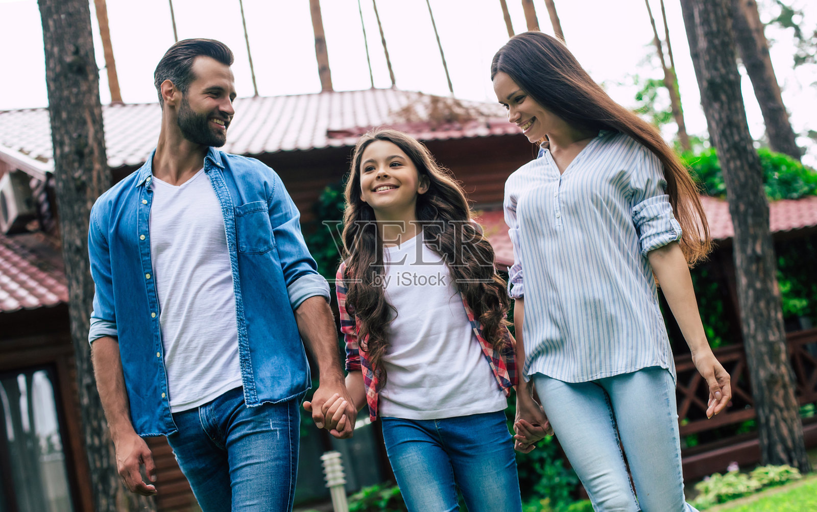 年轻快乐的现代父母带着一个可爱的女儿在户外散步，手牵手在郊区的房子背景。照片摄影图片