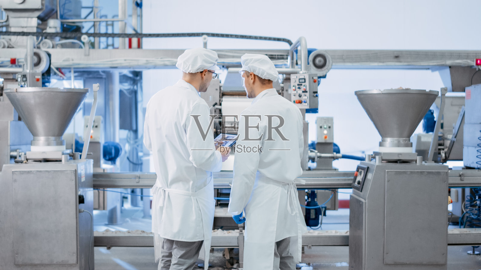 两个年轻的食品工厂员工讨论与工作有关的事情。男性技术员或质量经理使用平板电脑工作。他们戴着白色的卫生帽和工作袍。照片摄影图片