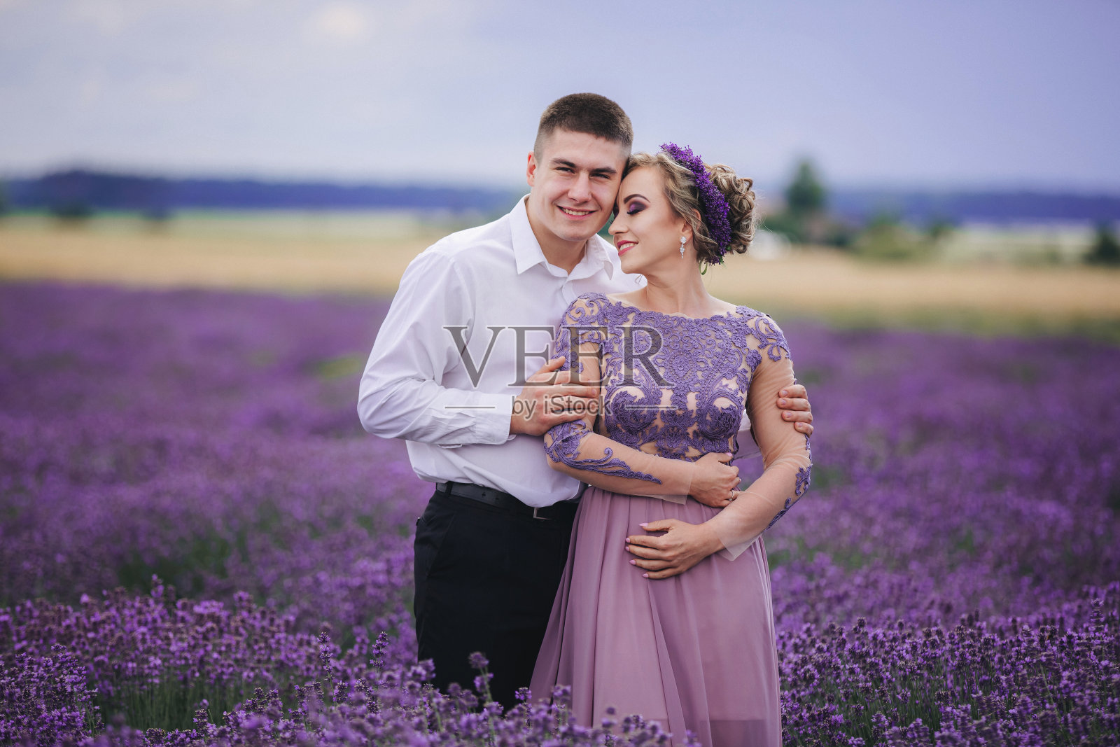 在夏天阴天的薰衣草地里，一对相爱的年轻夫妇拥抱着散步。穿着奢华的紫色衣服和发型的女孩照片摄影图片