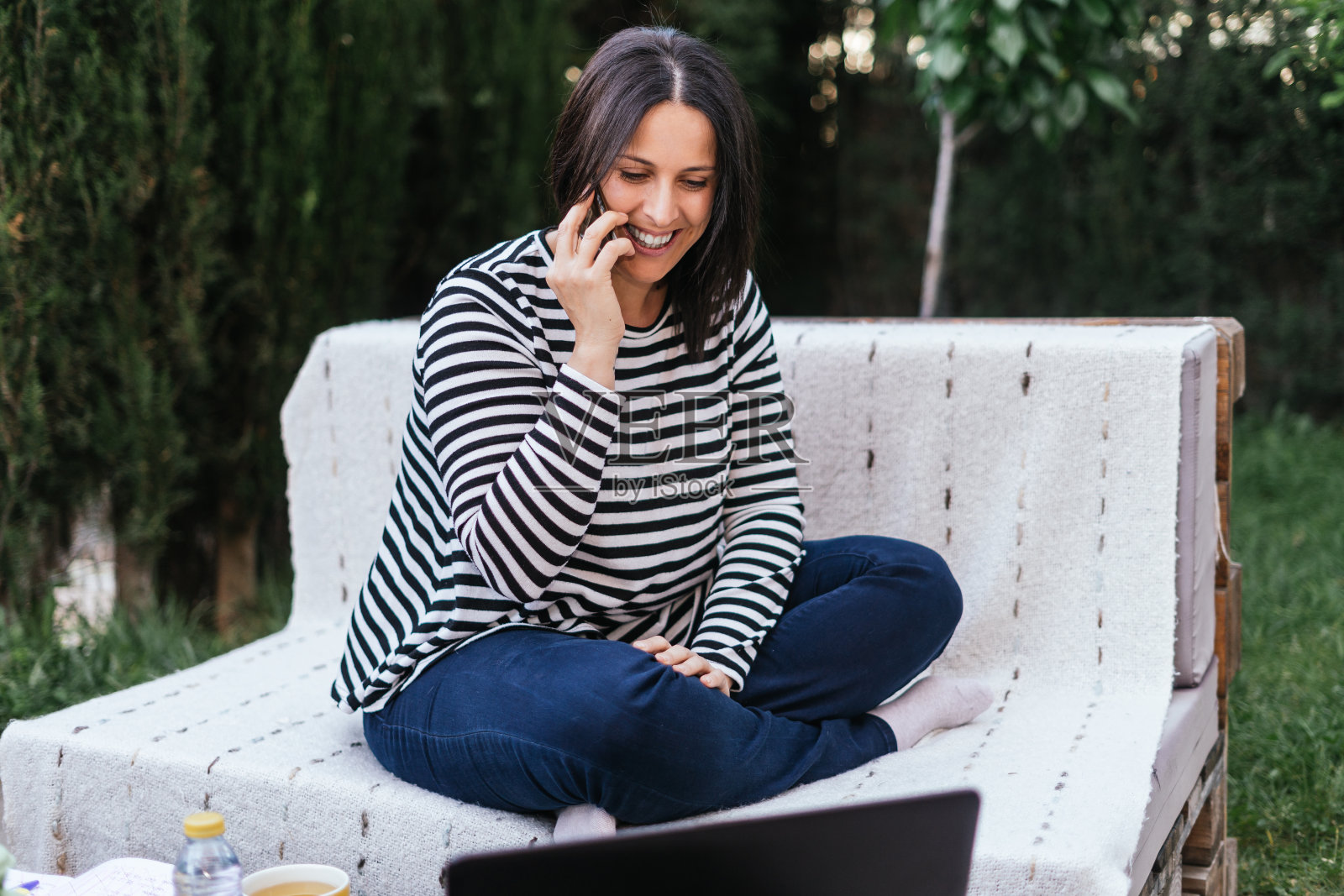 一位褐发女性在自家花园里用笔记本电脑远程工作，一边打电话一边微笑。她穿着深色牛仔裤和一件蓝白条纹毛衣。照片摄影图片