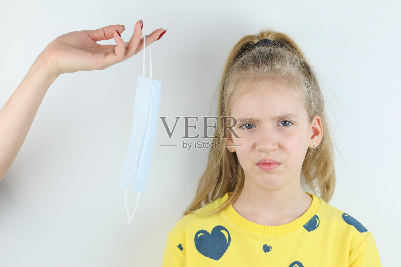 漂亮的孩子，穿着黄色t恤的女孩不想和拒绝戴医疗保健保护面罩和母亲的手在白色背景上便于提取照片摄影图片