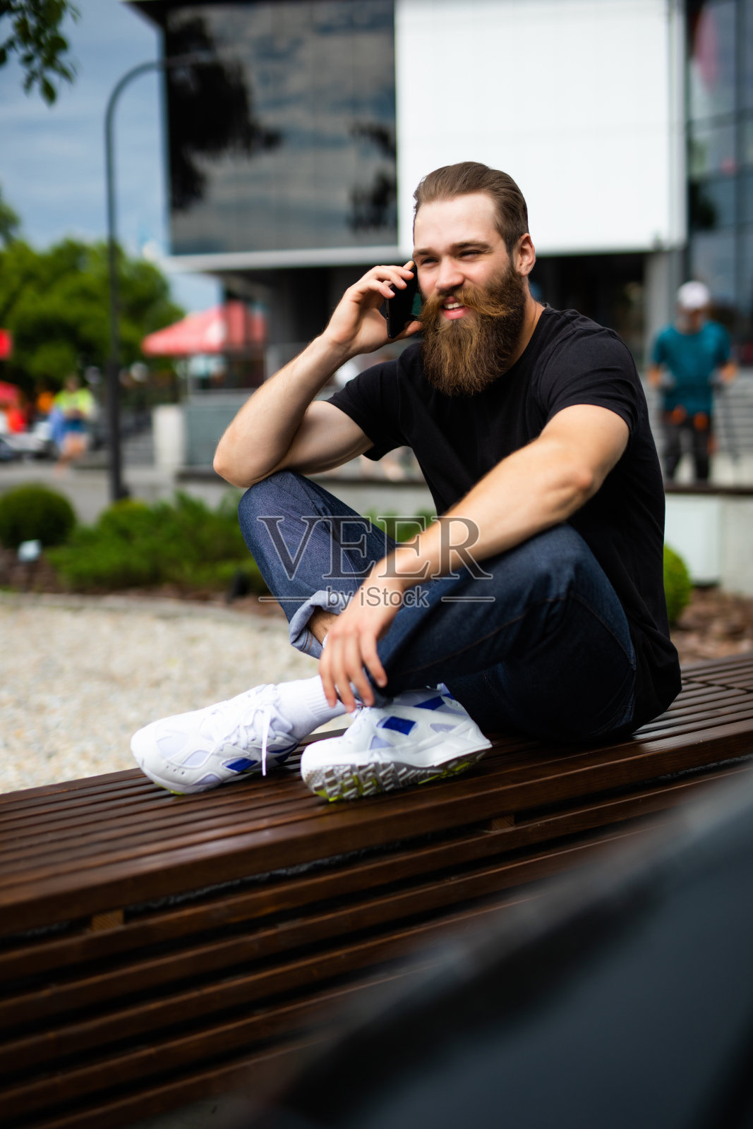一个蓄着胡须的野蛮男子坐在户外的长椅上，手里拿着手机。照片摄影图片