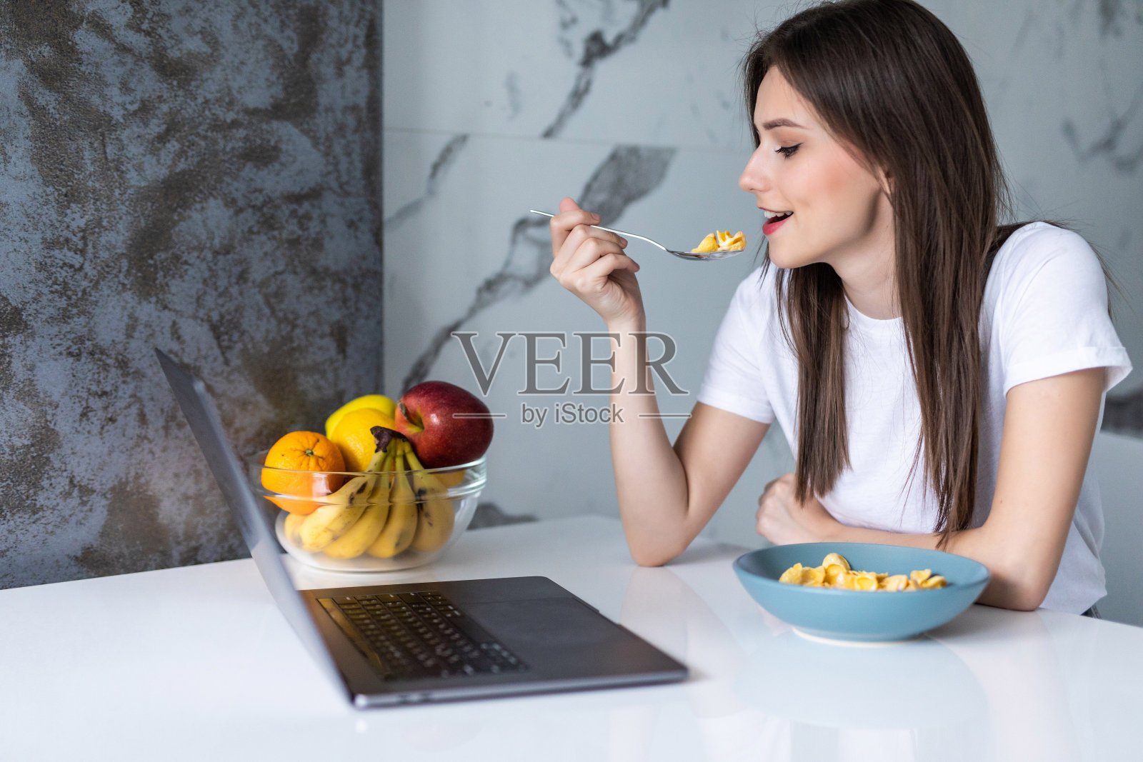 年轻女子在厨房用笔记本电脑吃早餐。穿着睡衣用笔记本电脑吃早餐的年轻女子。近距离照片摄影图片