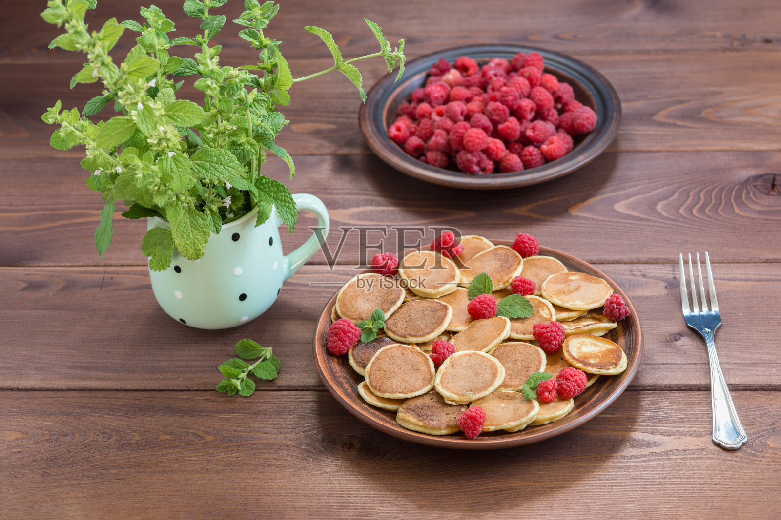 煎饼麦片和红色成熟的新鲜覆盆子放在一个棕色的盘子里。深色木桌上的马克杯里有一堆梅丽莎照片摄影图片