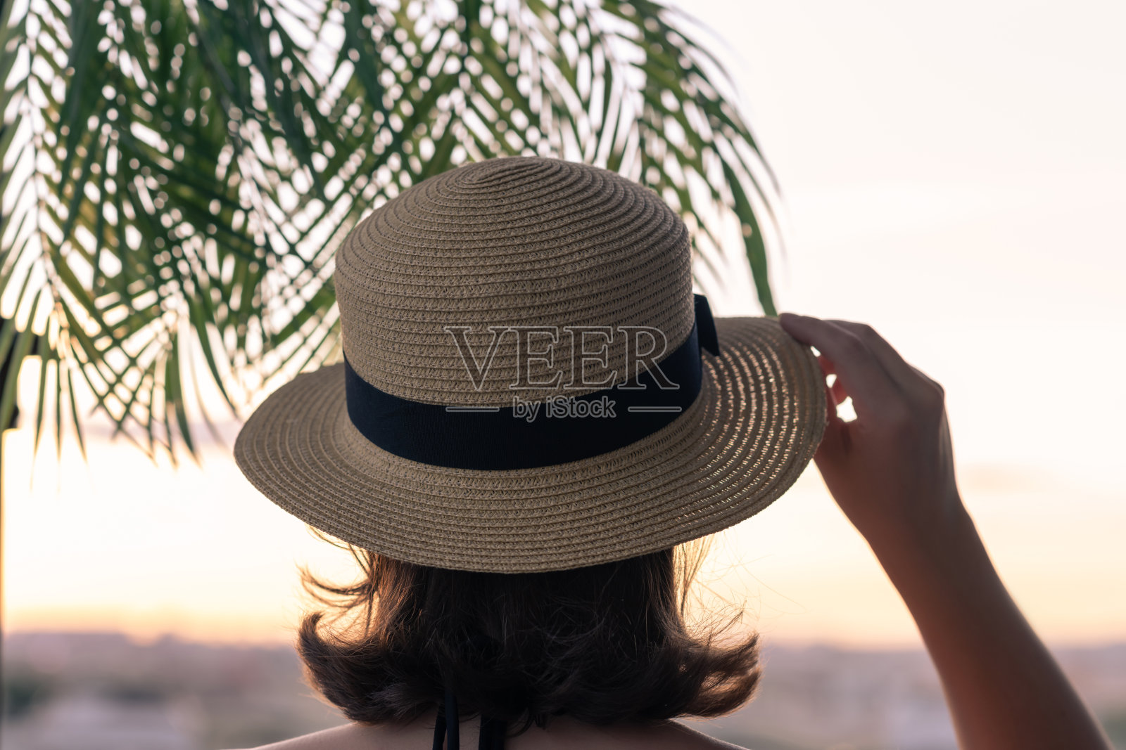 一个美丽的女孩戴着草帽背对着大海的背景在棕榈树的树枝。日落海滩。夏季和自由的概念。照片摄影图片