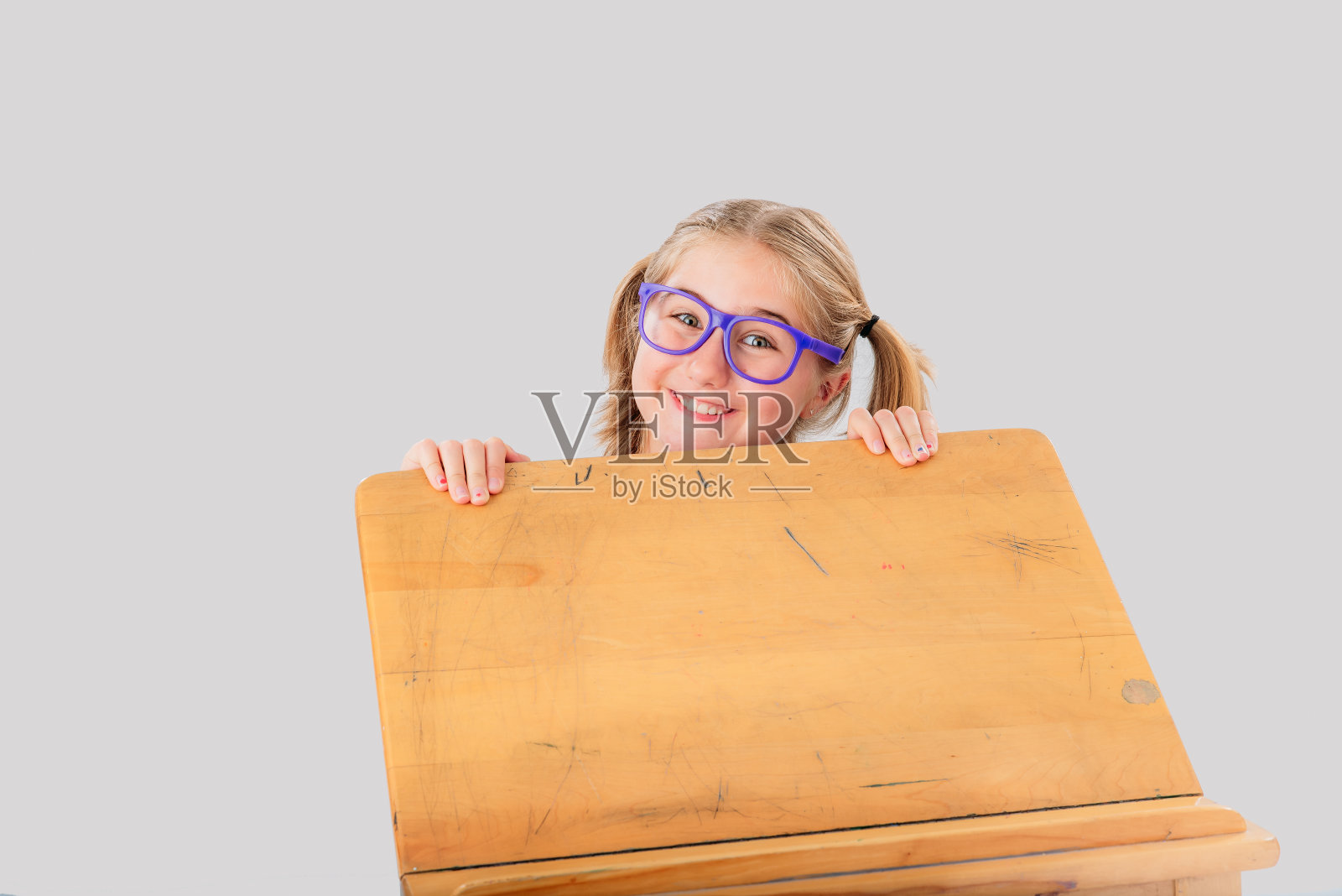 一个微笑的九岁小女孩藏在她的桌子后面，她戴着眼镜，扎着两个马尾辫。照片摄影图片